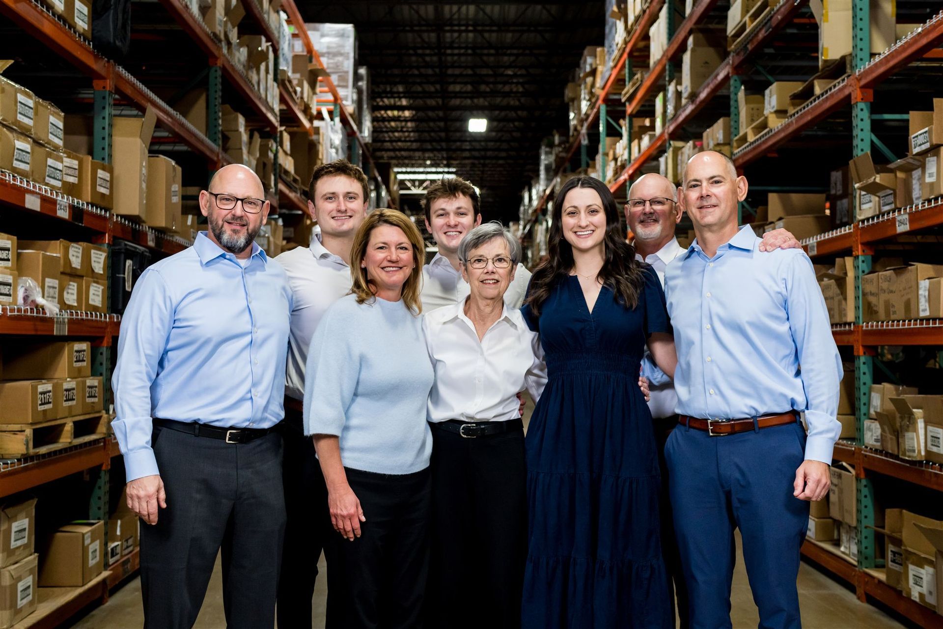 Group of seven people standing in a warehouse aisle with shelves of boxes, smiling at the camera