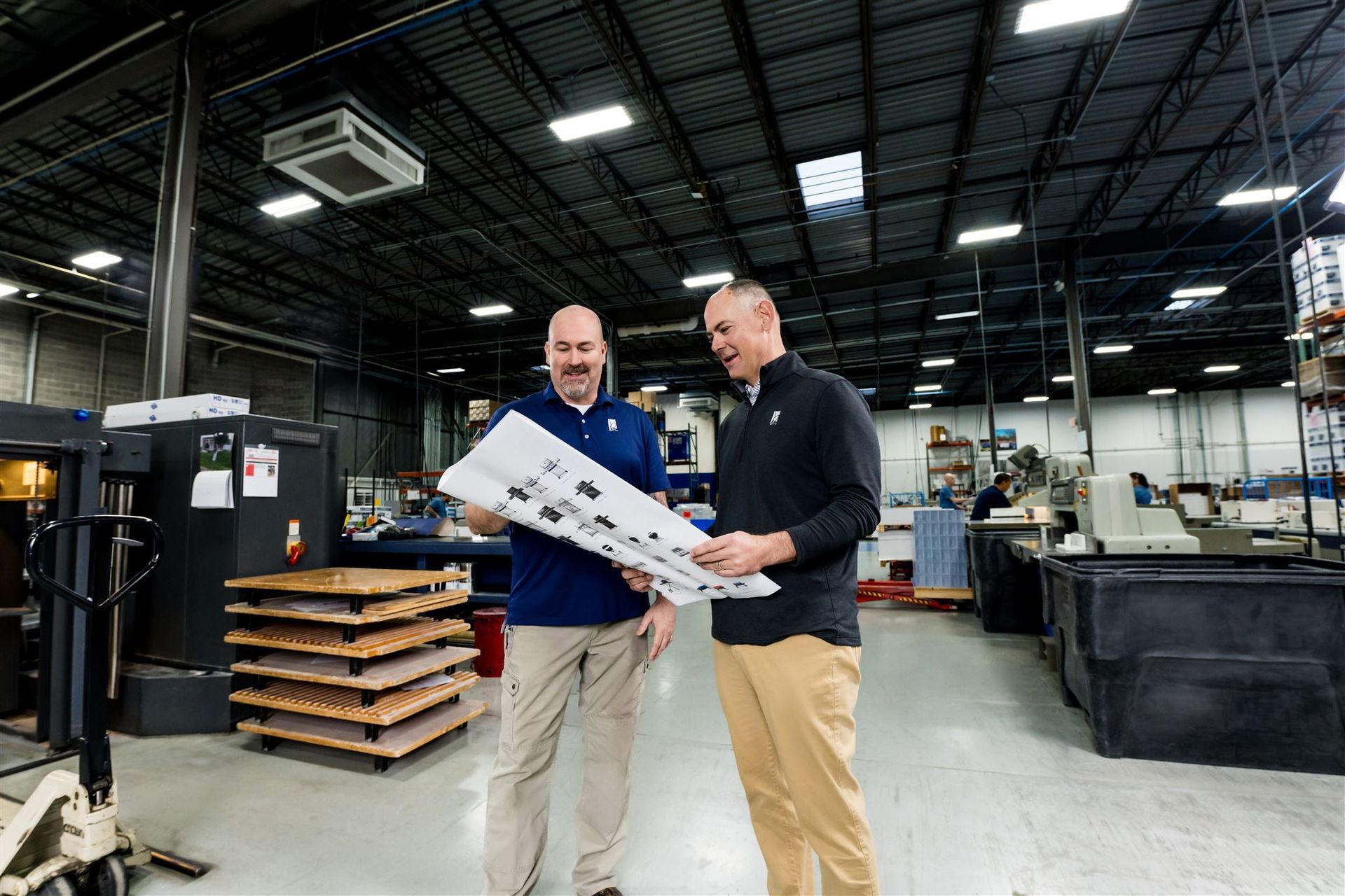 Two men in a warehouse holding a large printed sheet, with industrial equipment and pallets behind them