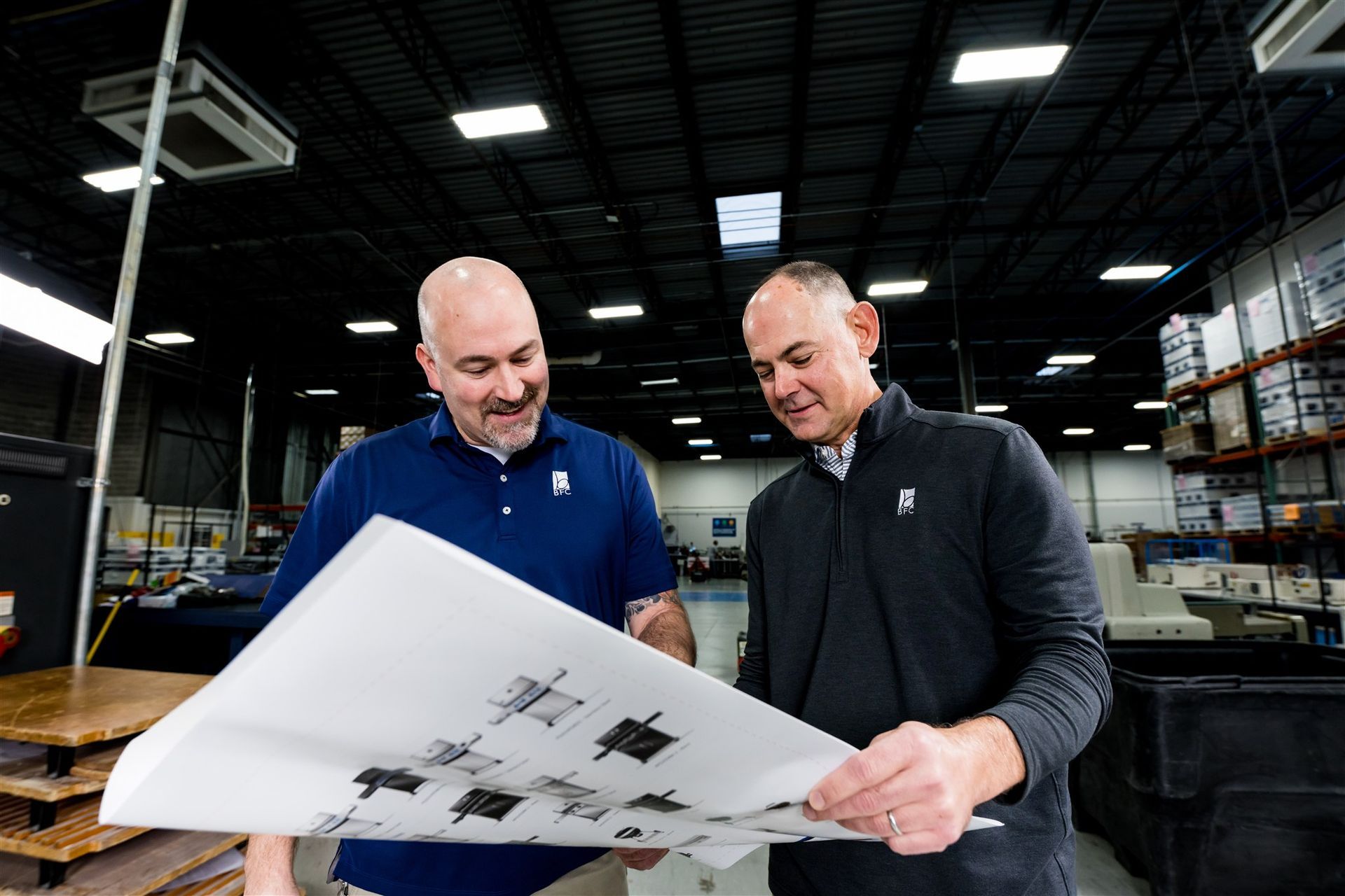Two men examining a large blueprint in a warehouse with shelves and overhead lights.