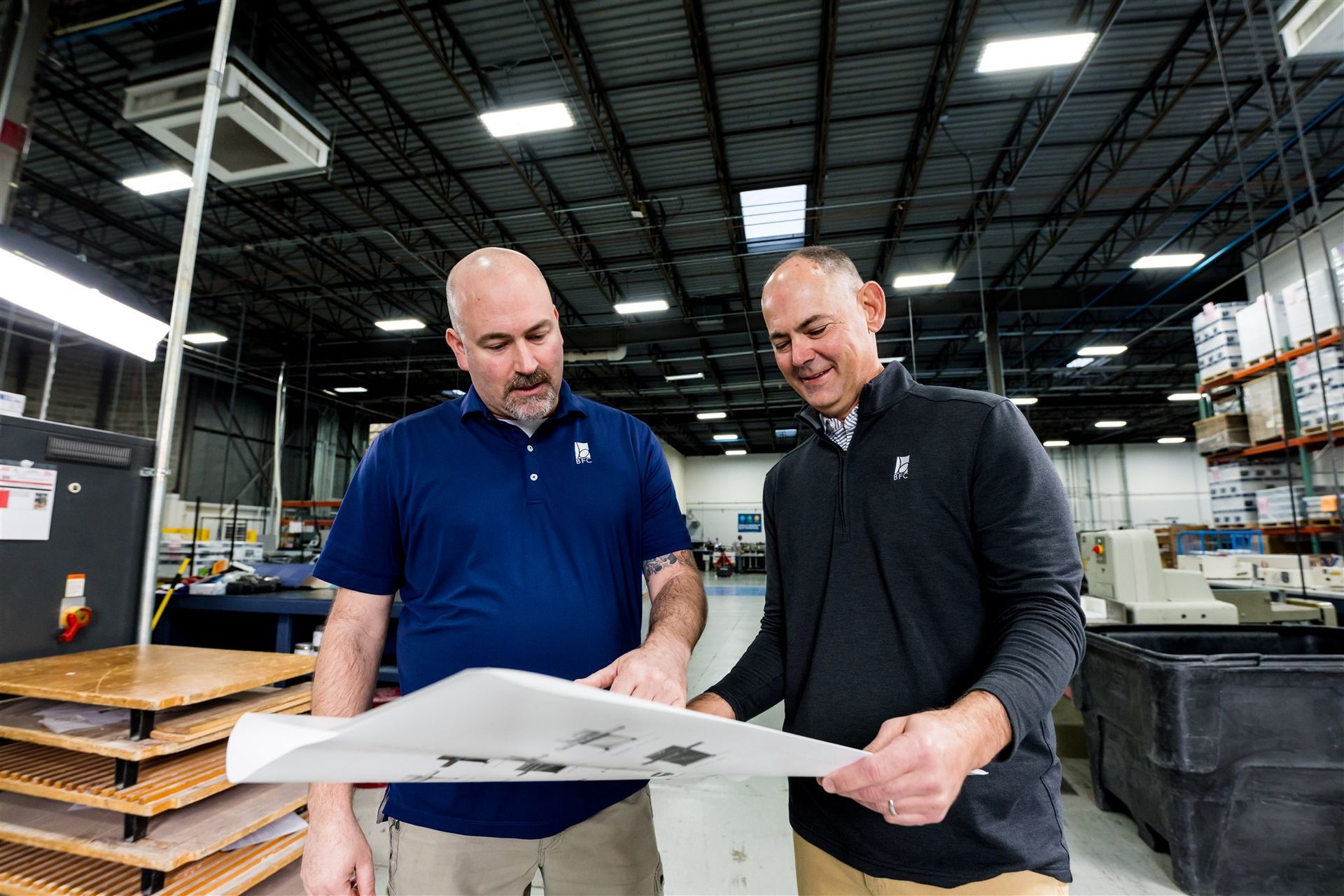 Two workers in a warehouse review a large printed plan together