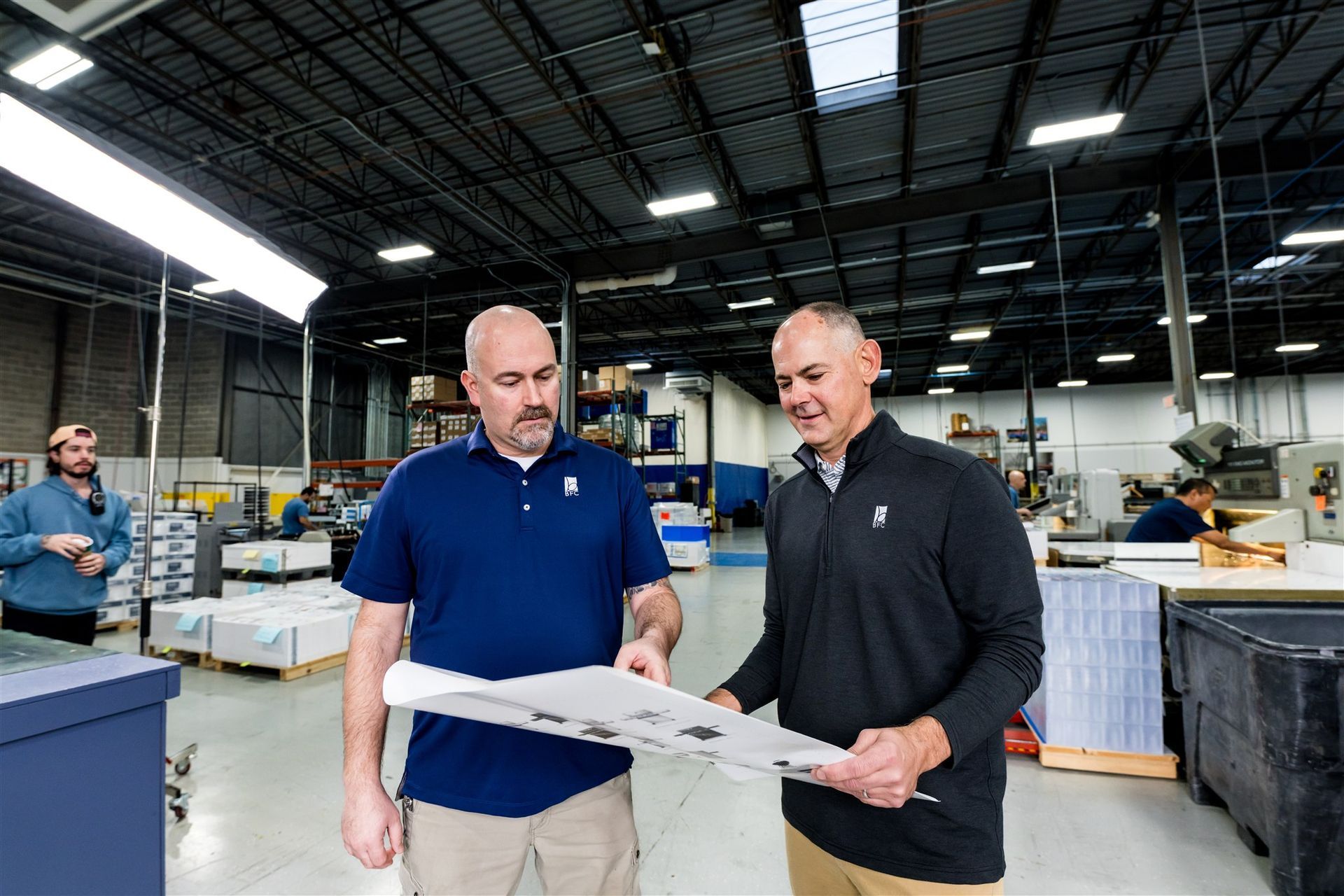 Two men reviewing a blueprint inside a warehouse with stacked materials and machinery in the background