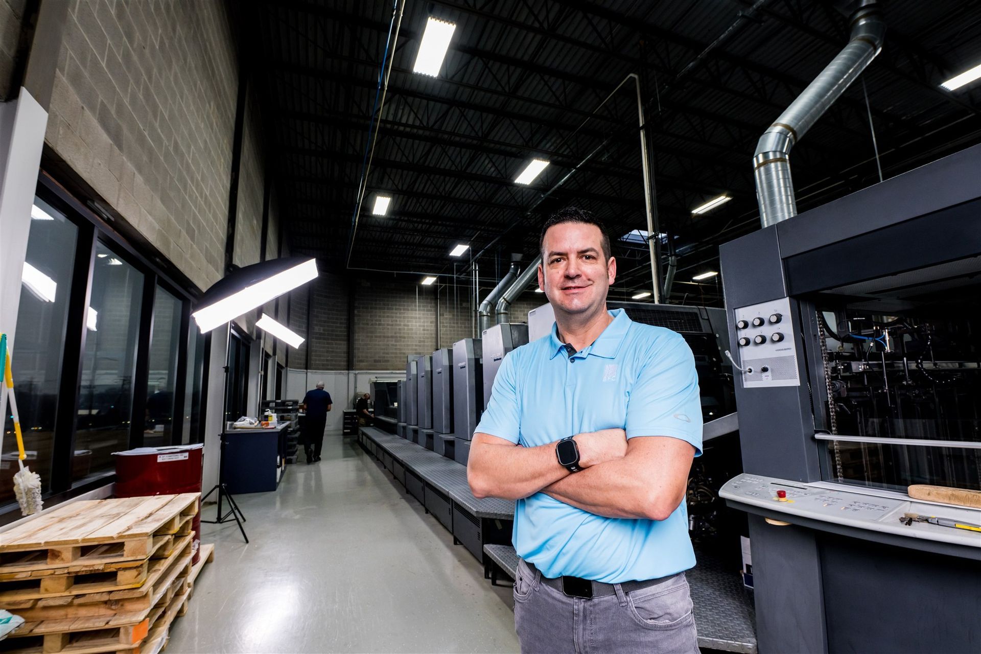 Man standing with arms crossed in a warehouse aisle, surrounded by industrial equipment and pallets.