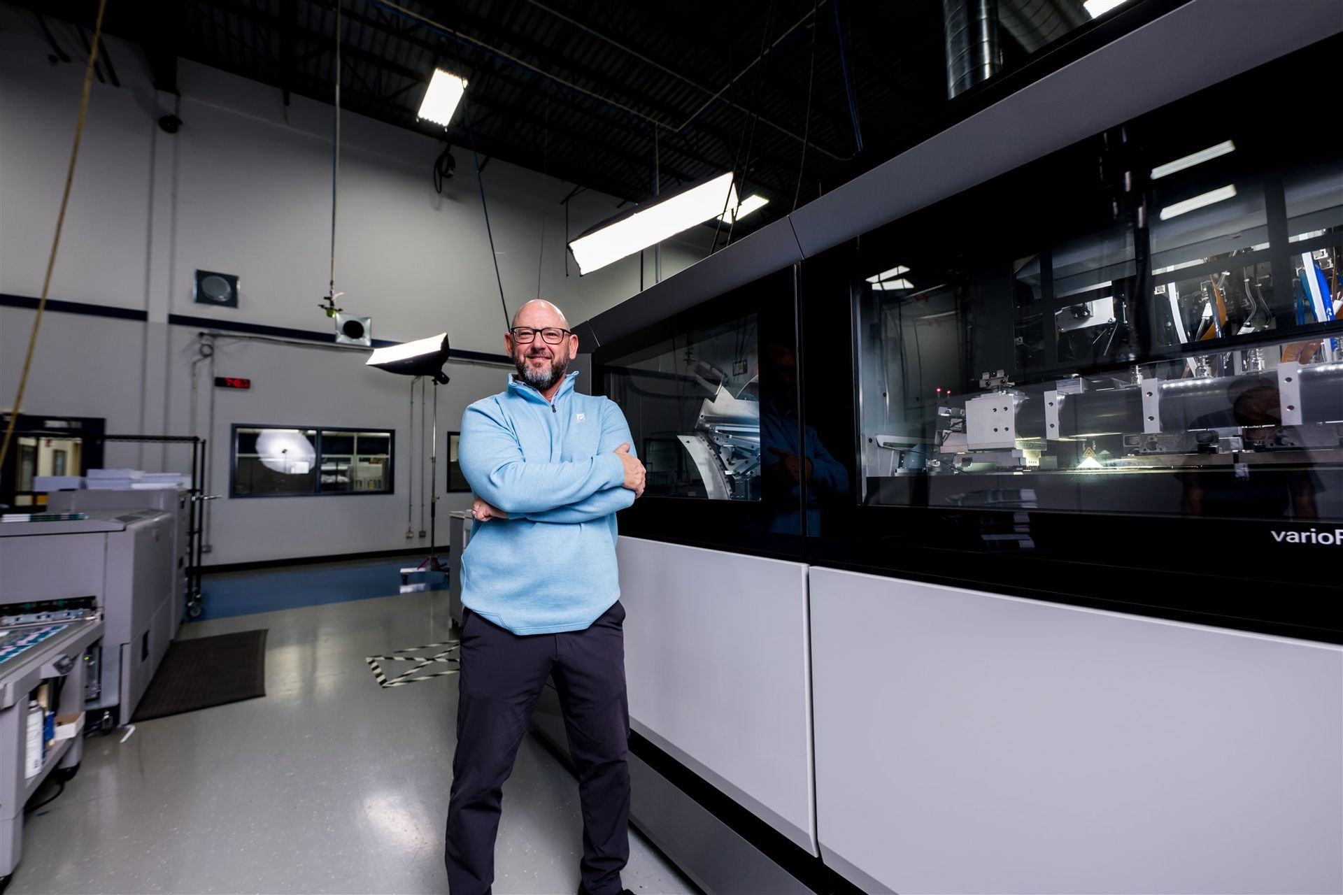 Man standing with arms crossed in an industrial workshop beside large machinery and glass panels