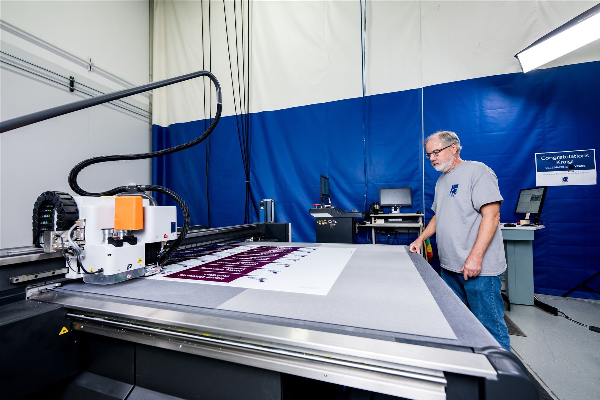 Man operating a large flatbed printer in a workshop with a blue wall and printing equipment