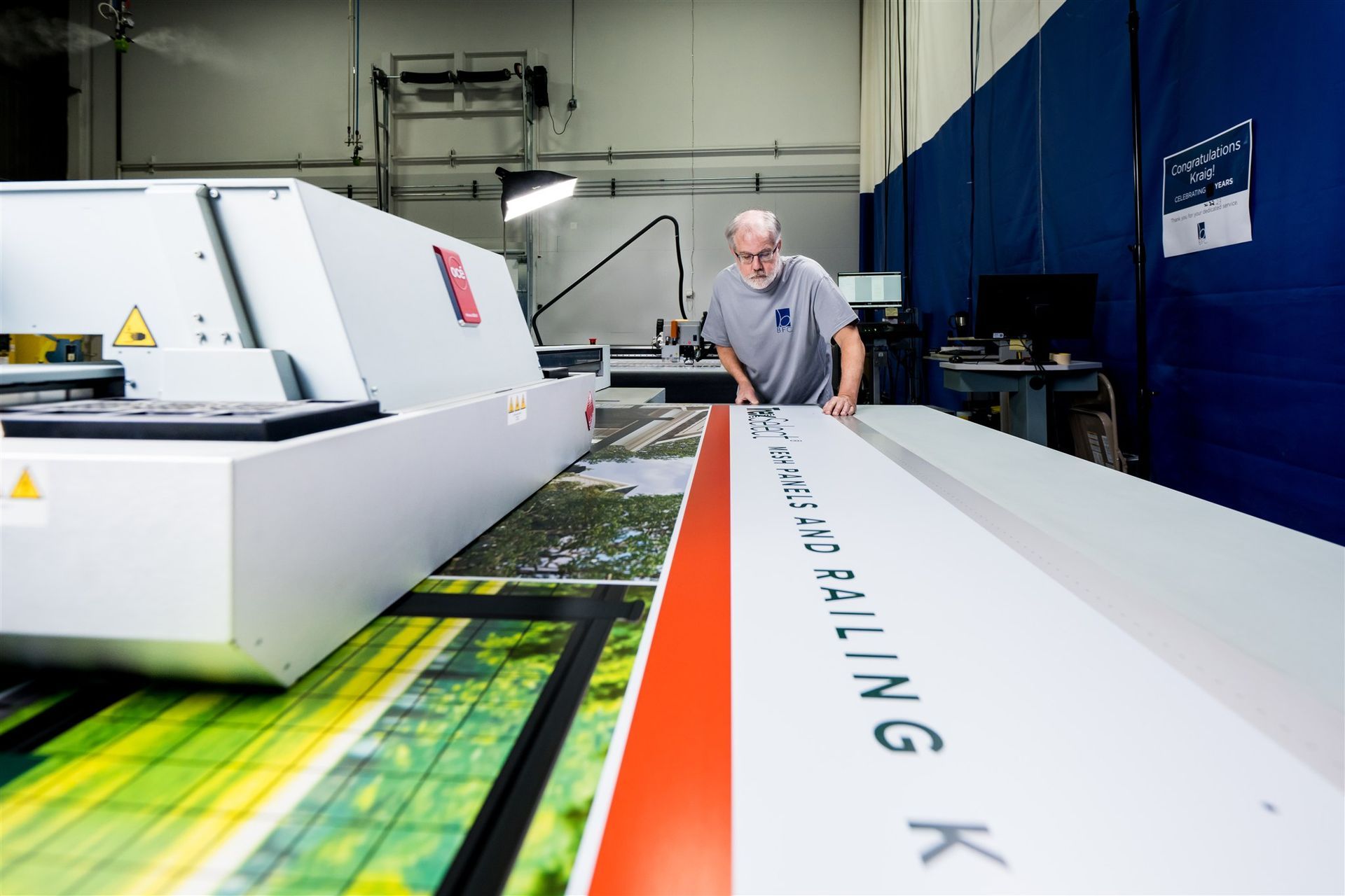 Worker inspecting large industrial printing machine in a factory, with white and orange panels.