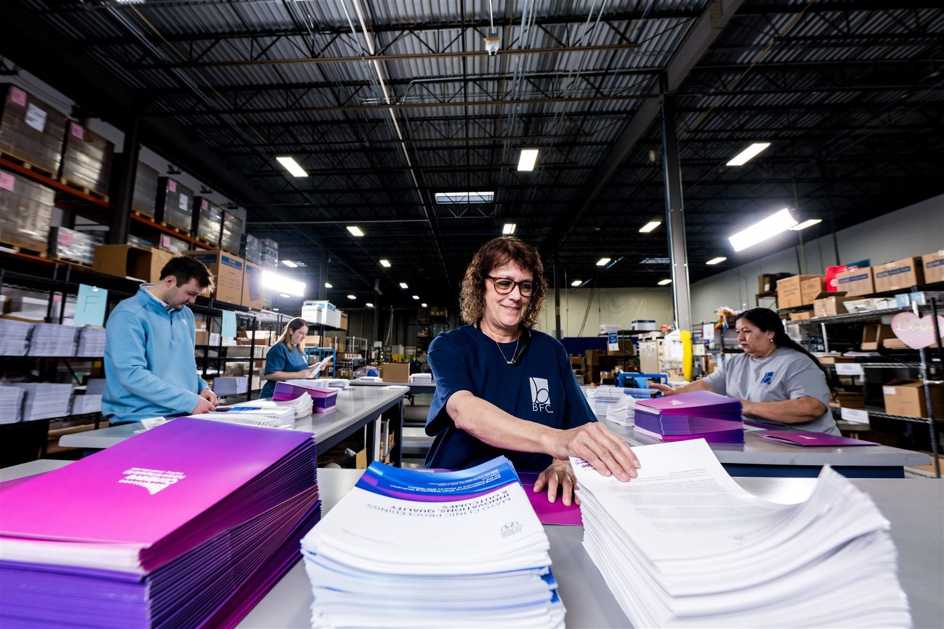 Workers sort stacks of printed paper in a large warehouse with high shelves and overhead lights.