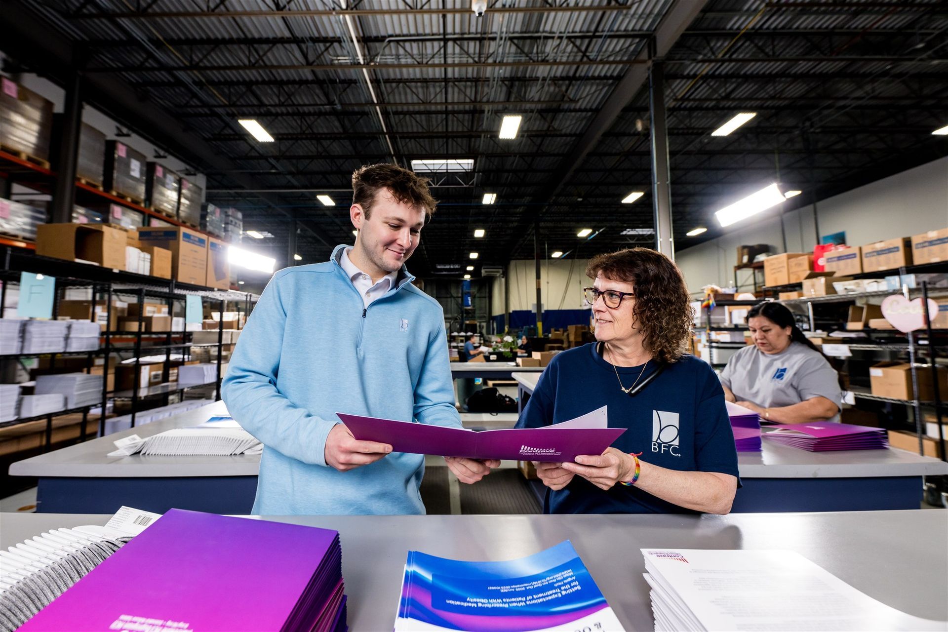 Two workers reviewing a purple folder in a warehouse with shelves of boxes and supplies.