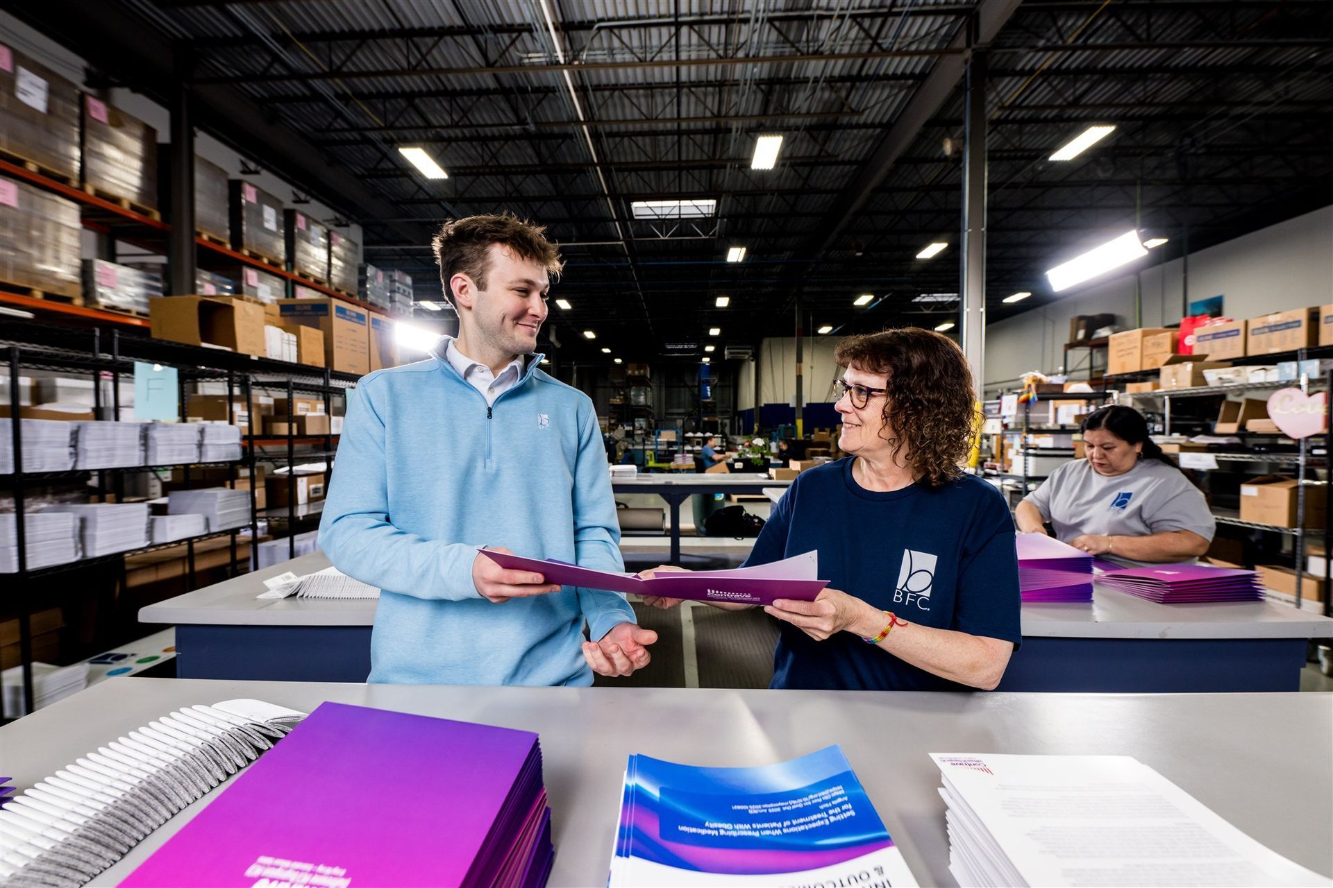 Workers sorting packages in a warehouse, with a man handing a pink folder to a woman at a table
