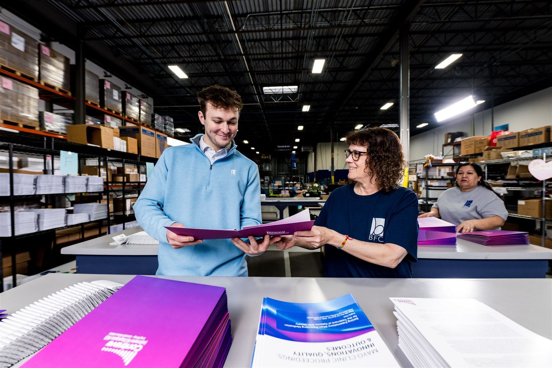 Workers packing folders in a warehouse, with stacks of boxes and paperwork on tables