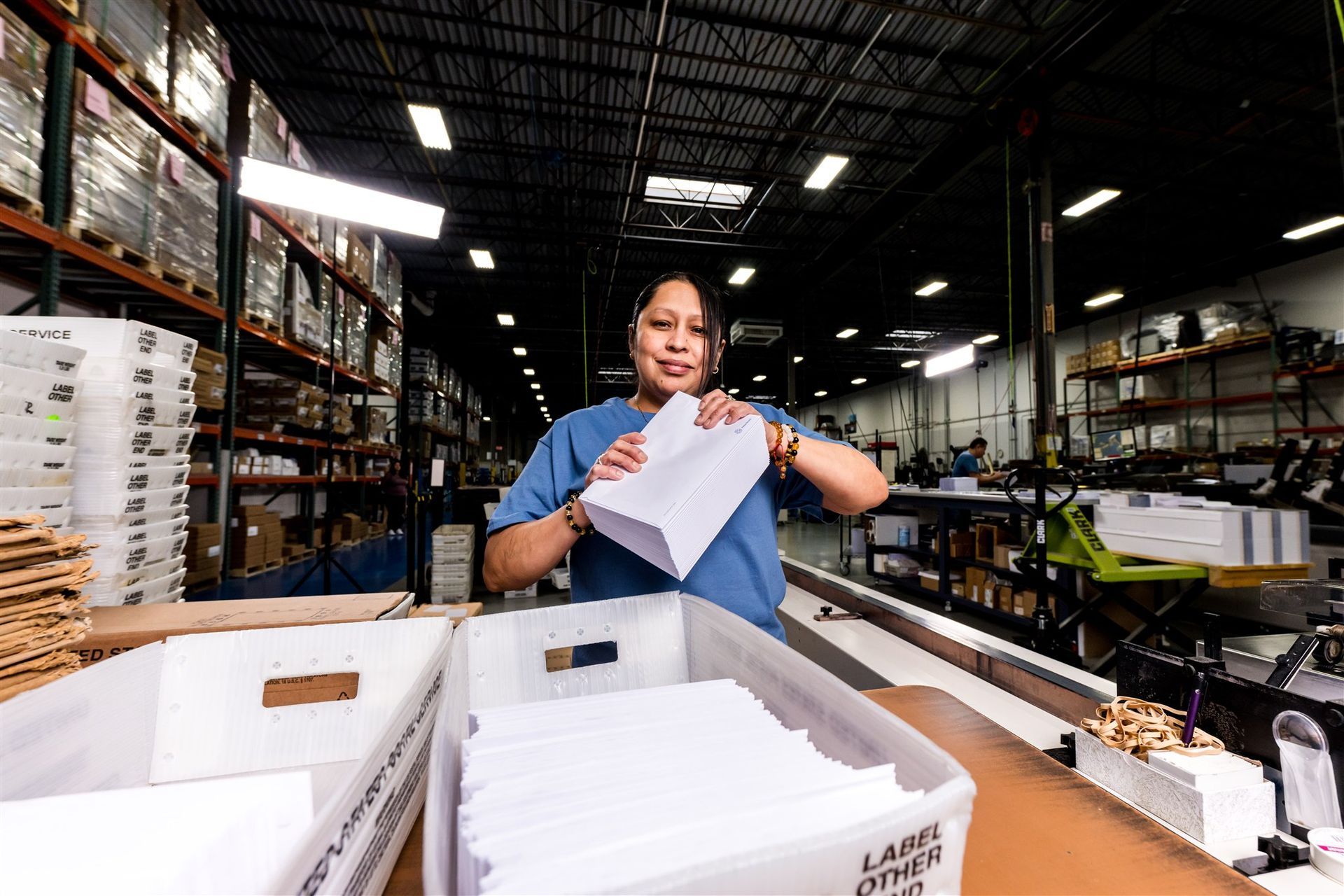 Worker sorting papers in a warehouse aisle with stacks of boxes and shelves in the background