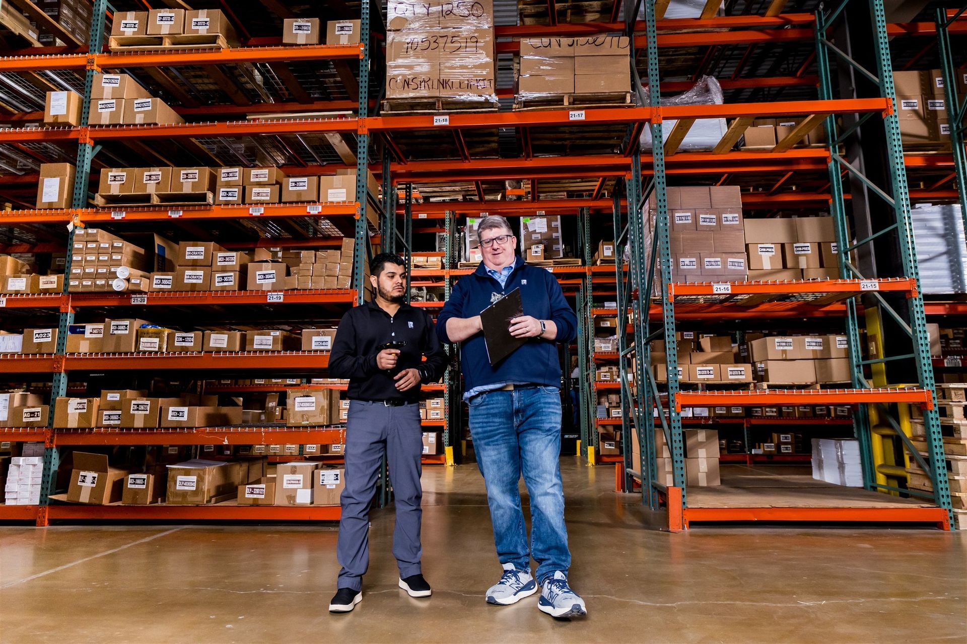 Two people stand in a warehouse aisle beside tall orange shelving stacked with boxes.