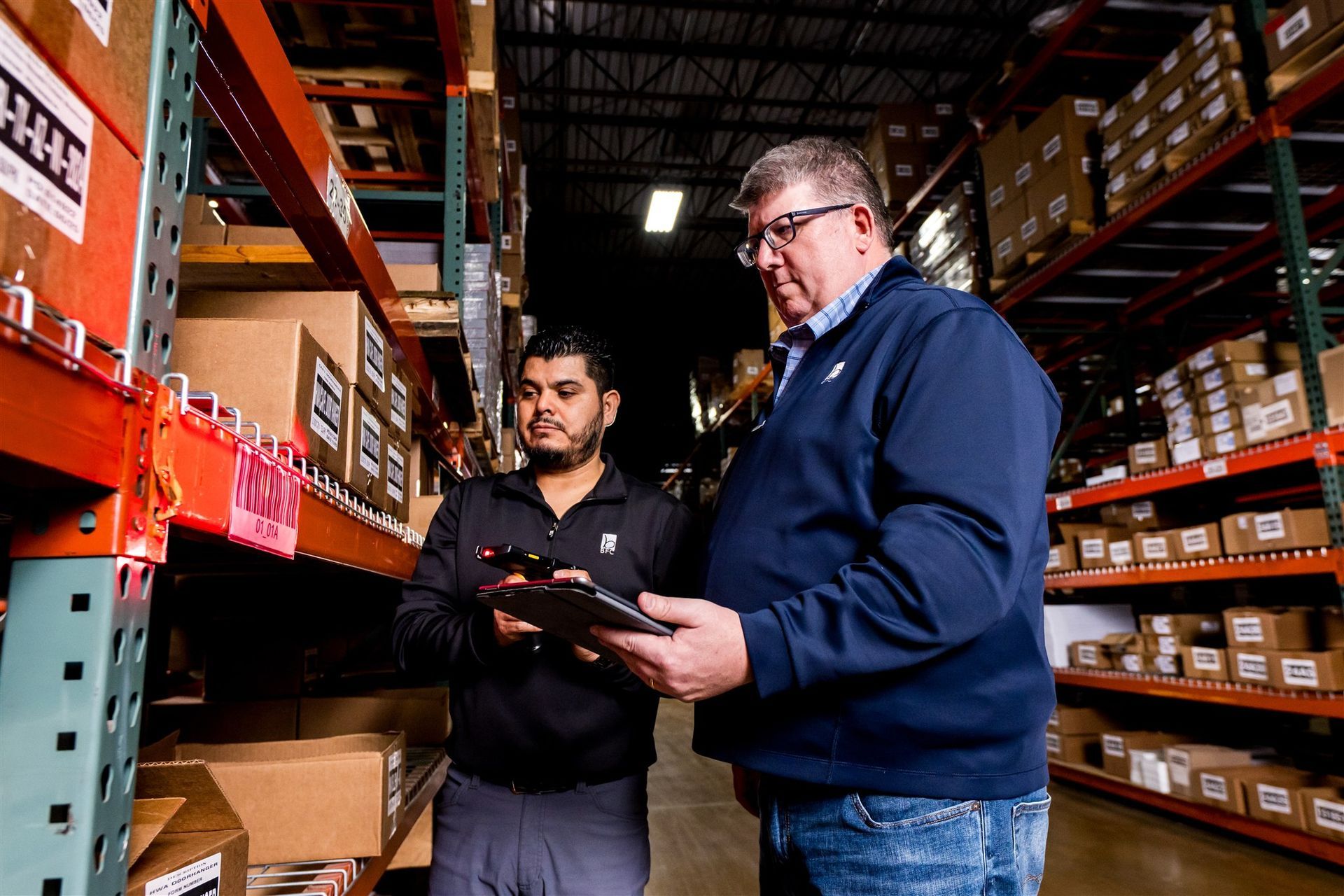 Two people reviewing a tablet in a warehouse aisle with shelves of boxed inventory