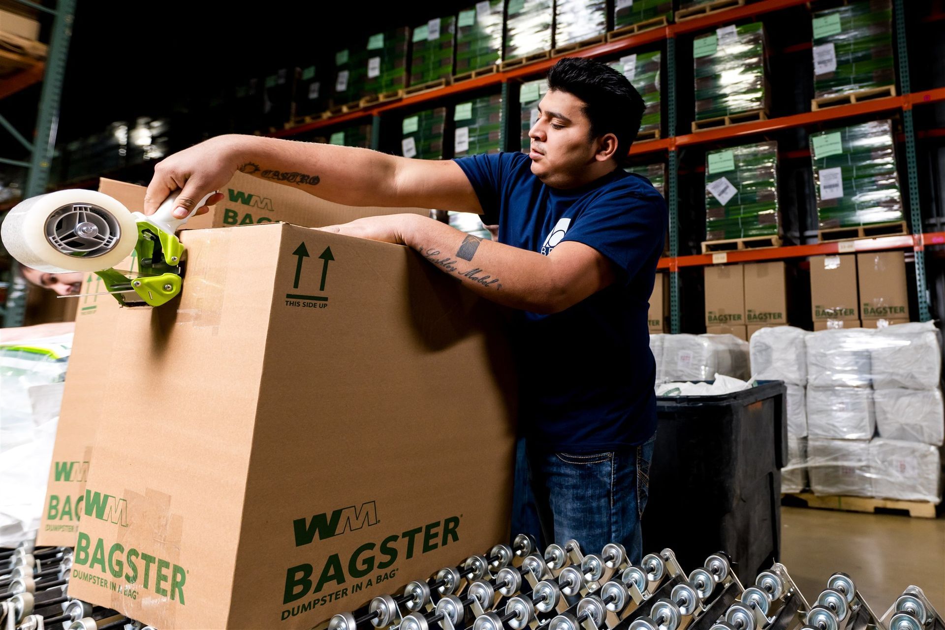 Worker stacking cans in a warehouse beside a large cardboard box labeled BAGS TER