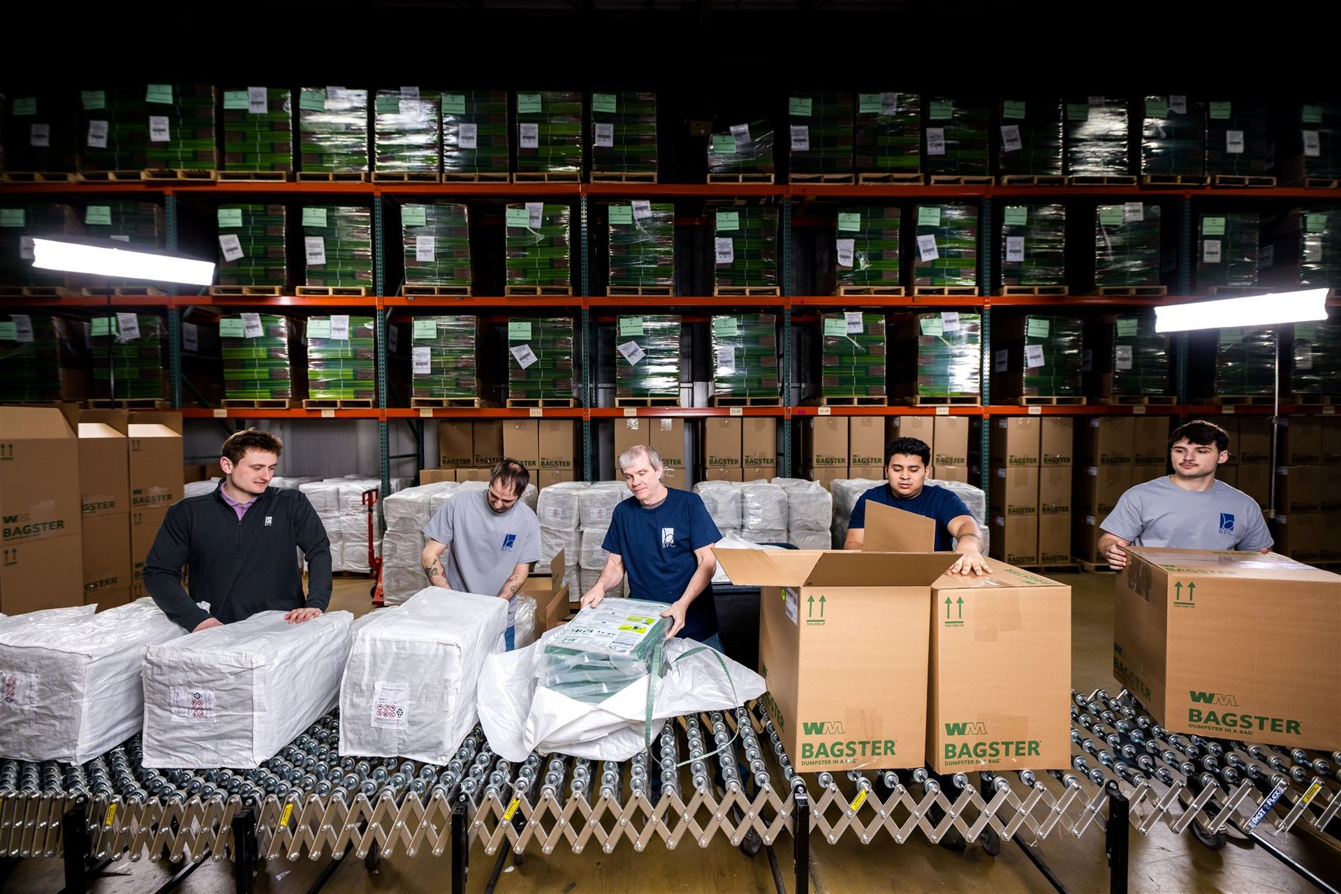 Workers packing pallets of boxed goods in a warehouse with shelves stacked high behind them