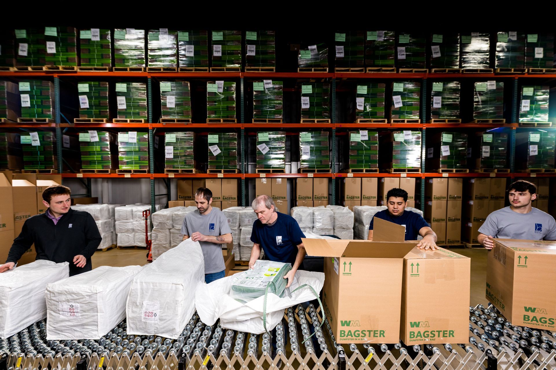 Workers packaging goods in a warehouse, surrounded by stacked boxes and shelves of inventory.