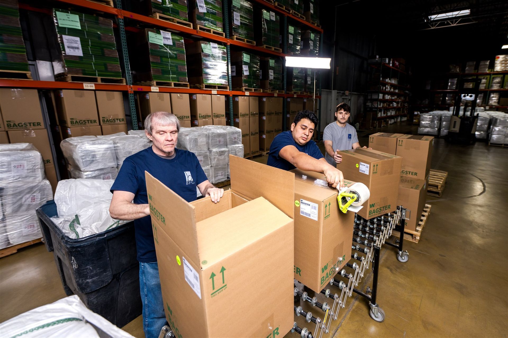 Workers packing boxes in a warehouse aisle with shelves and stacked pallets in the background