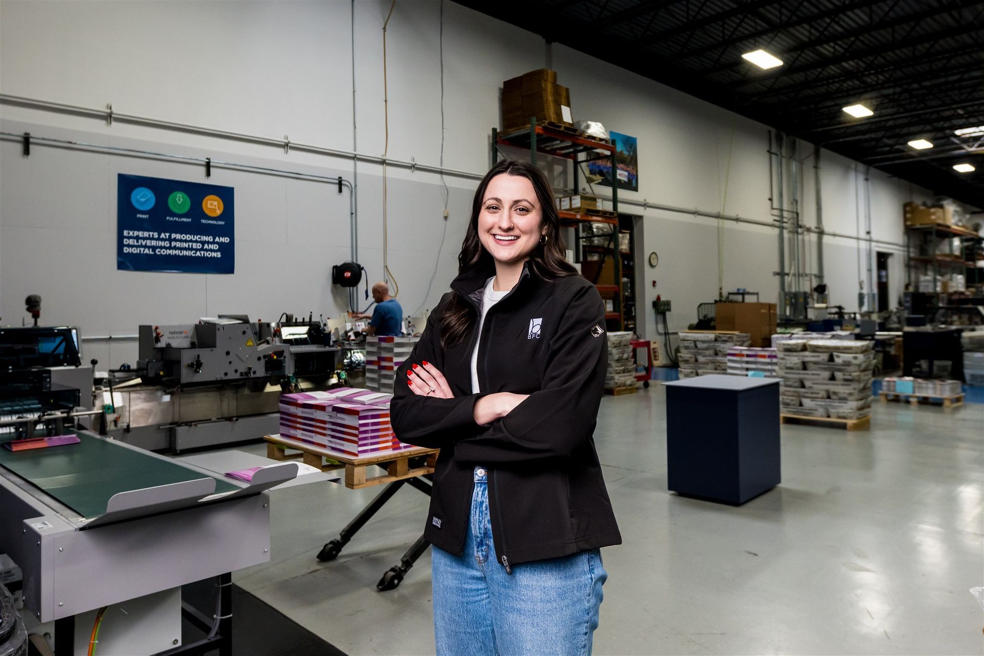 Smiling woman with arms crossed in a warehouse with shelves, equipment, and pallets in the background