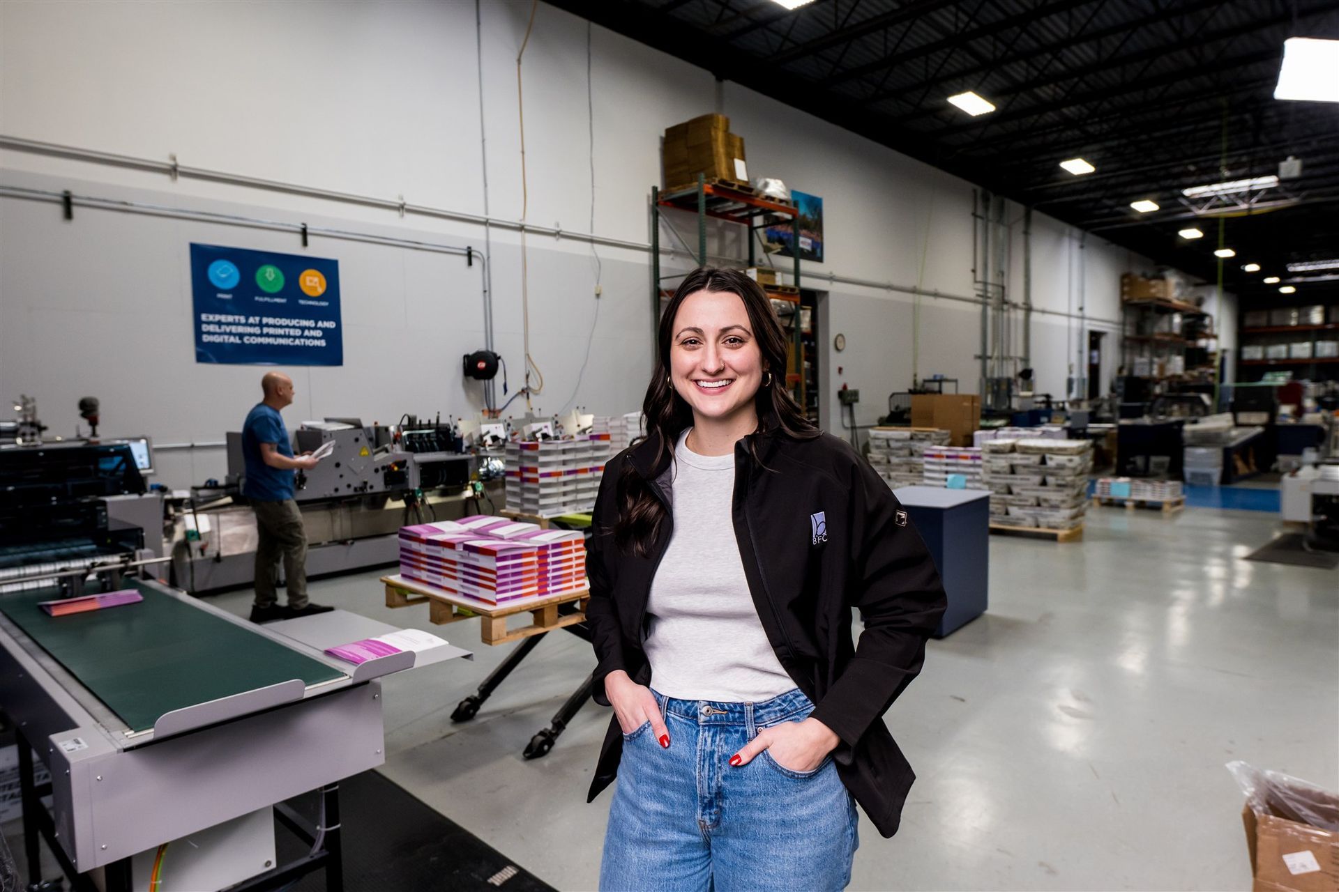 Smiling woman standing in a warehouse with industrial equipment and a conveyor belt behind her