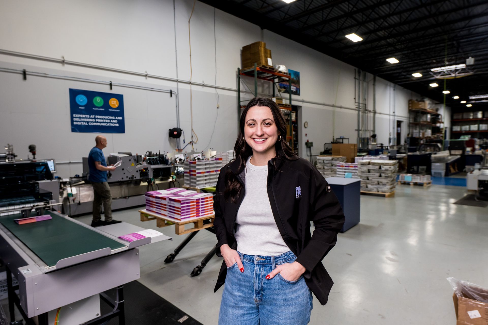 Smiling woman stands in a print shop with large machines and stacked materials in the background