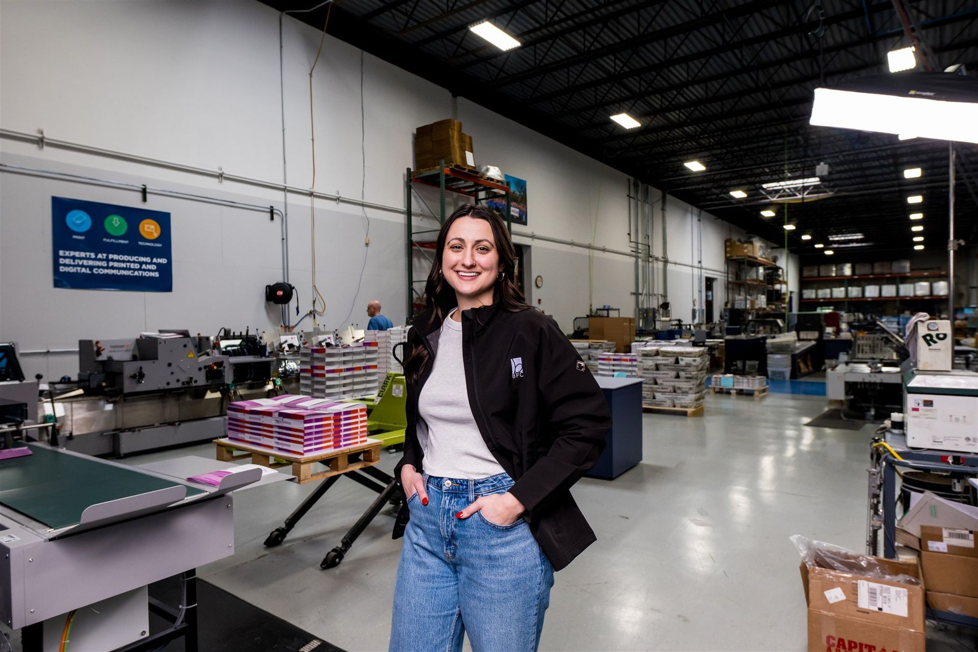 Woman smiling in a factory warehouse beside industrial equipment and stacked boxes