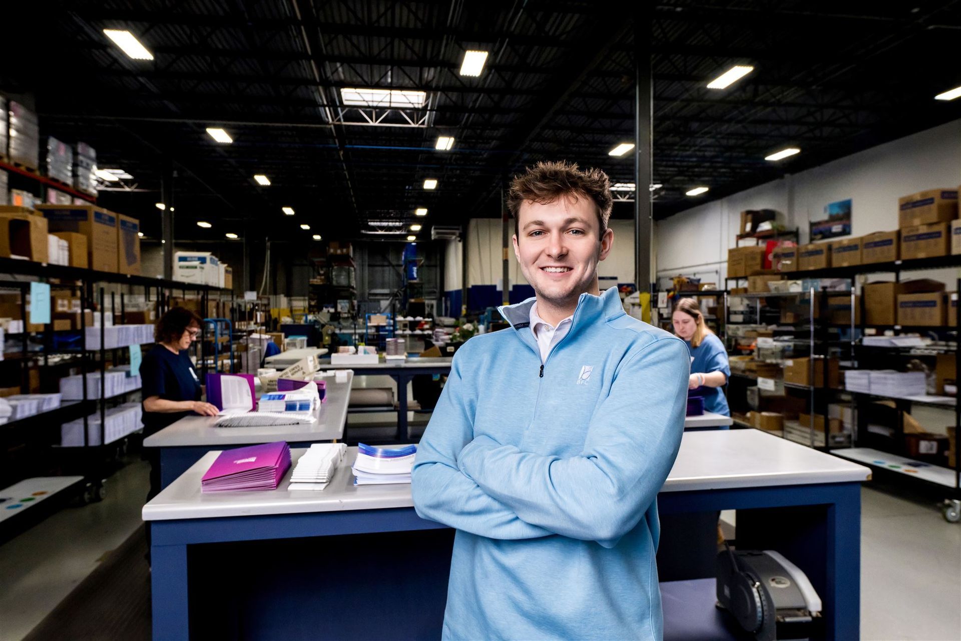 Person smiling with arms crossed in a warehouse fulfillment center, with shelves and workers behind.