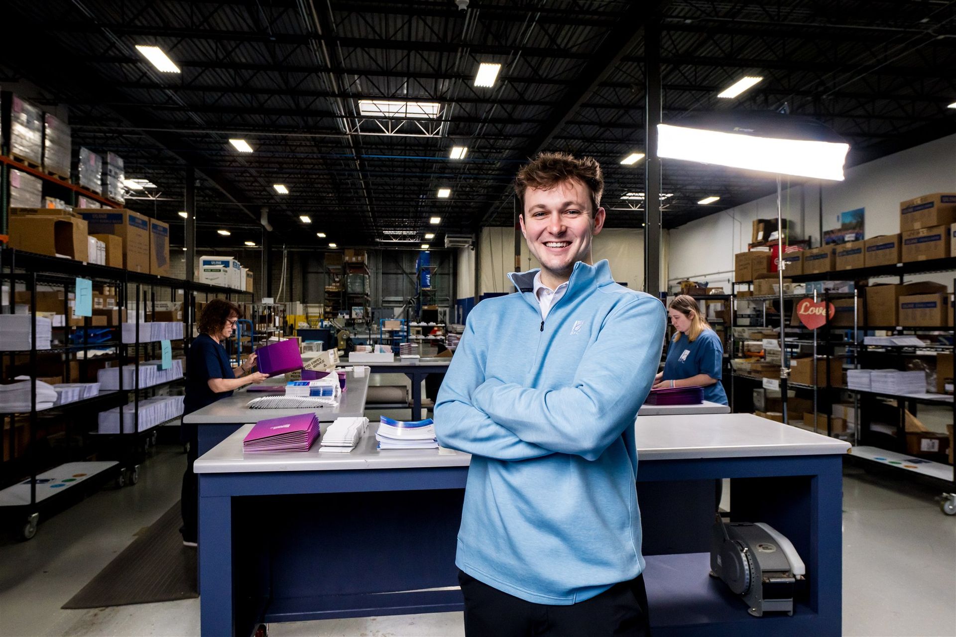 Smiling man in a blue shirt stands in a warehouse with shelves, boxes, and workers in the background