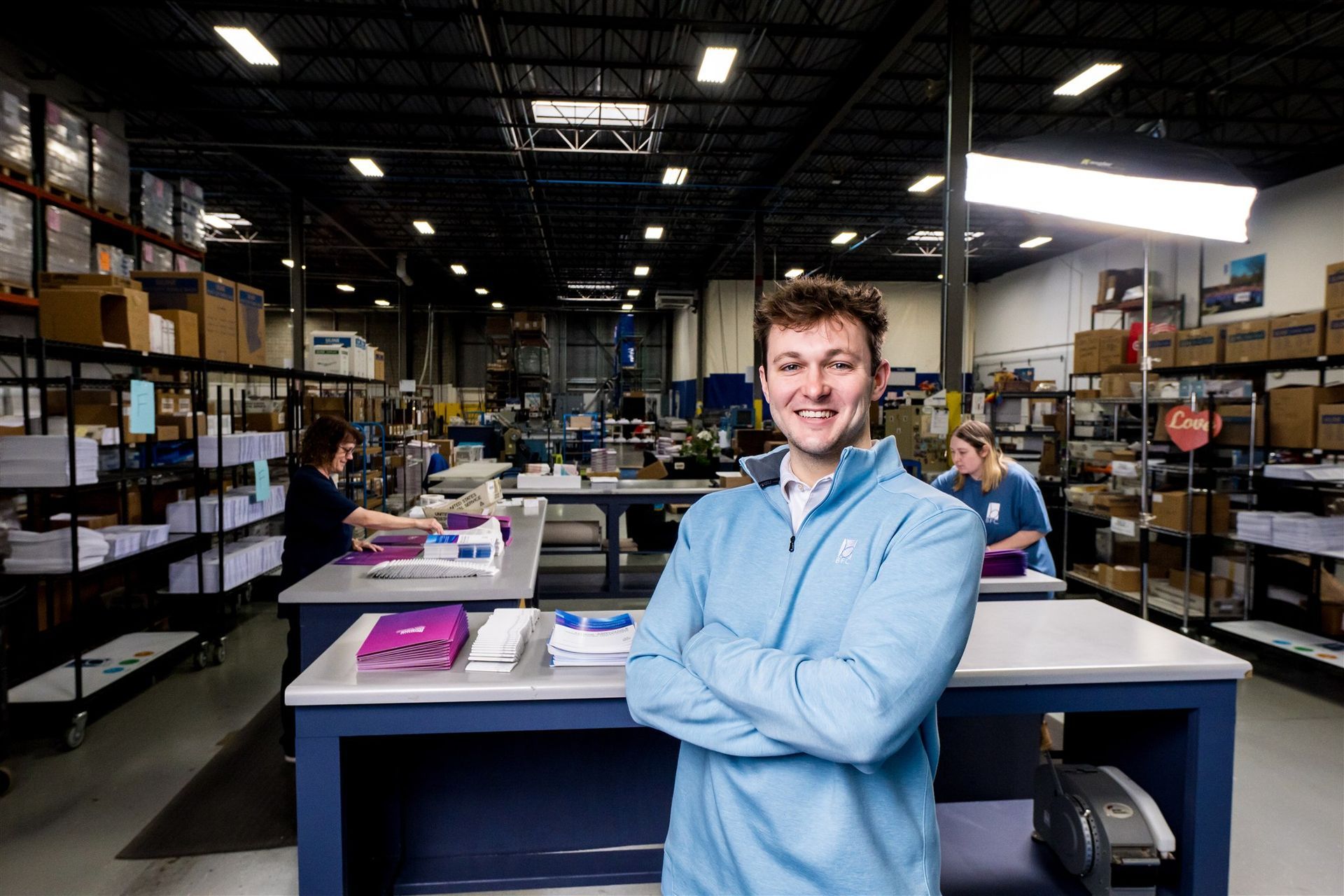Person in a blue lab coat smiling in a warehouse workspace with shelves and boxes in the background