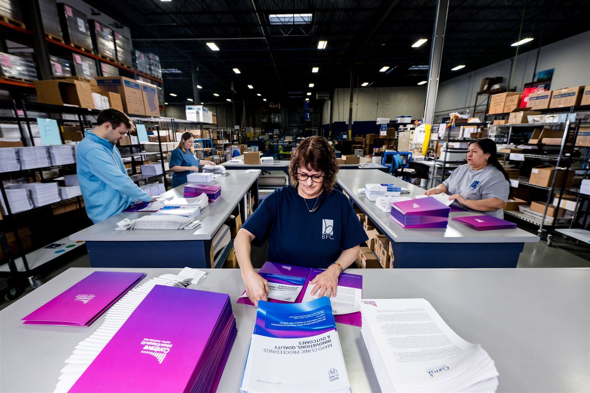 Workers packing items at assembly tables in a warehouse, with stacks of bright pink boxes.