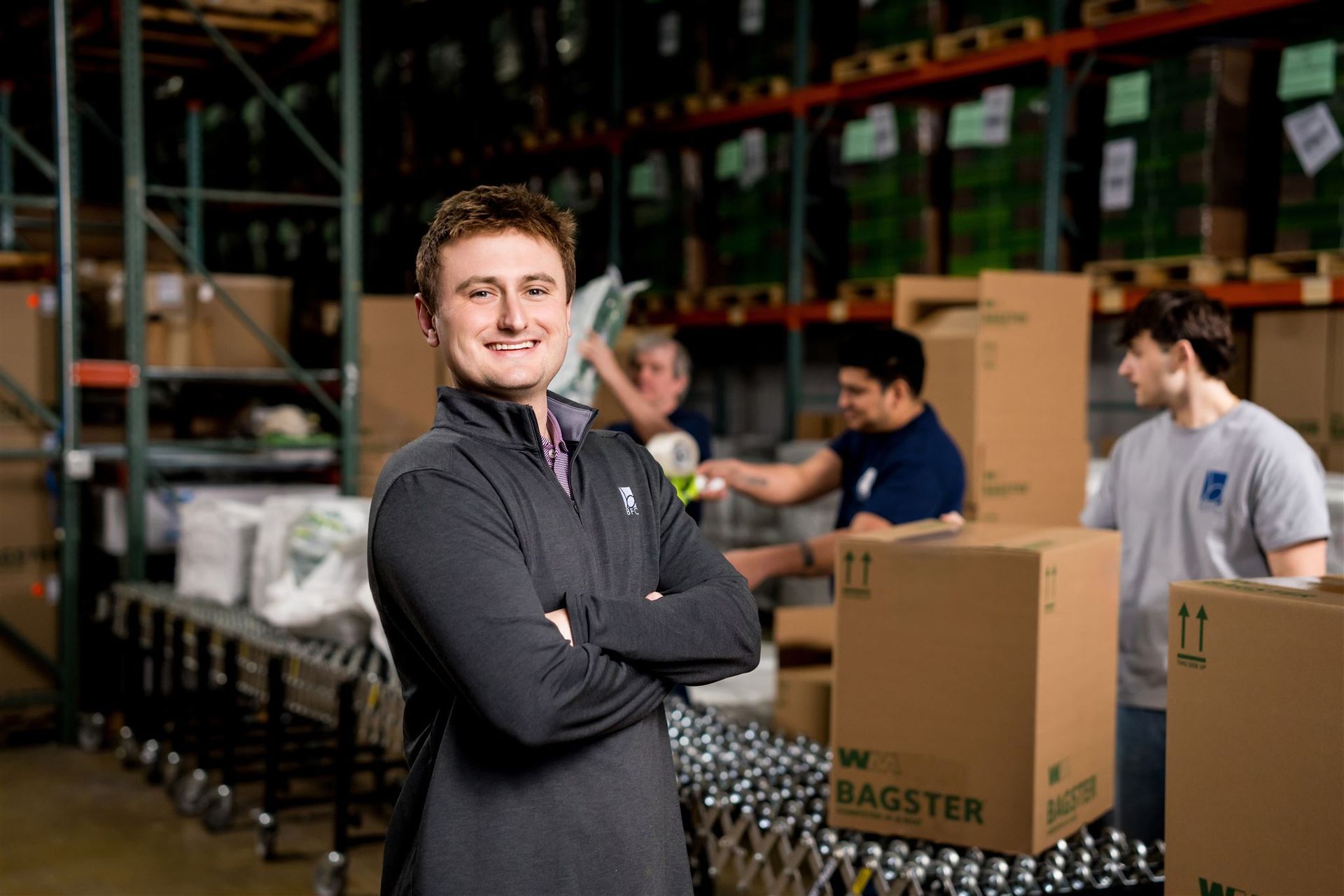 Warehouse staff standing by stacked boxes and bottles, with a man in a gray sweater smiling in the foreground