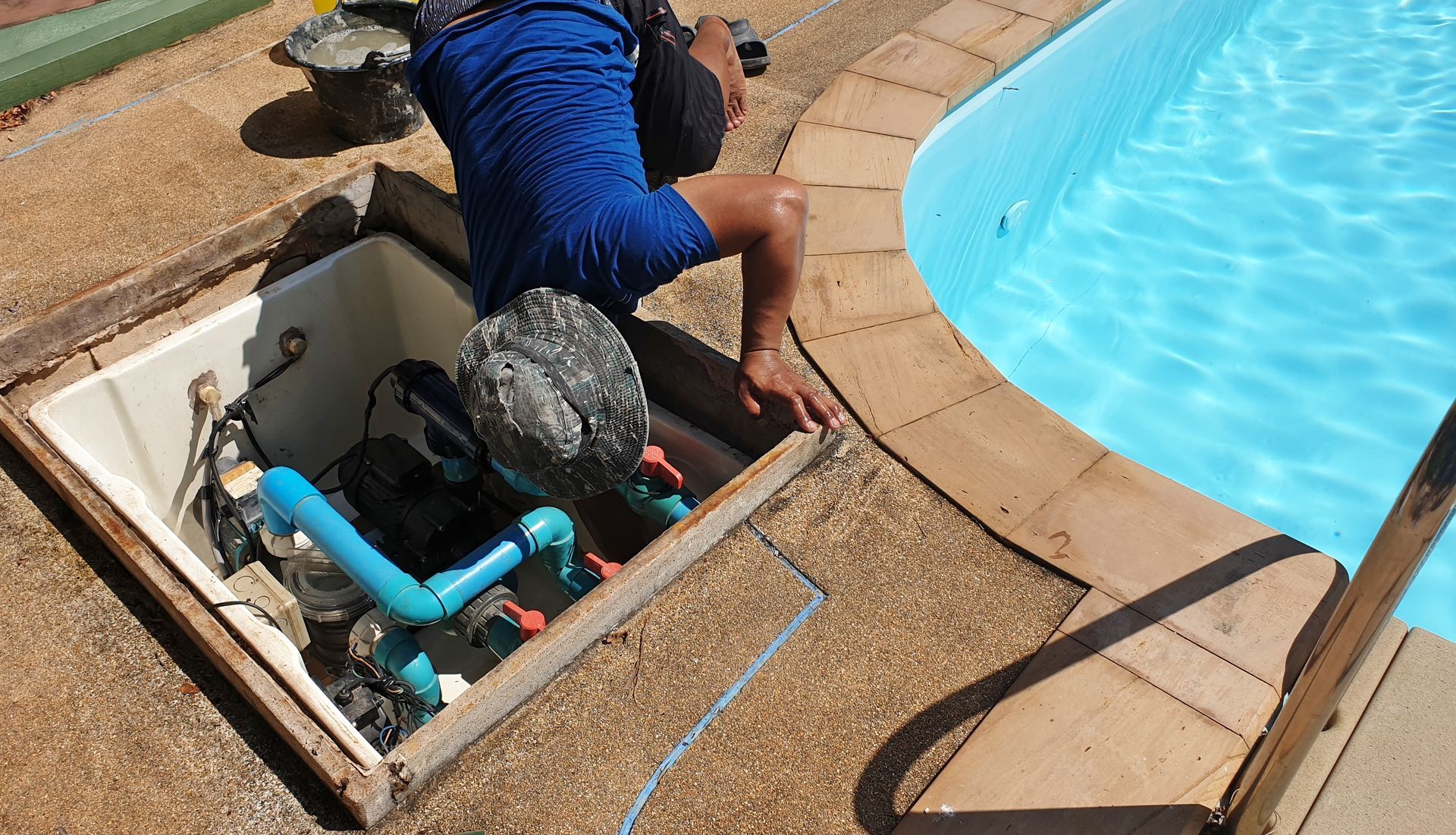 Person working on pool equipment beside a swimming pool, with a blue hose in an open hatch.