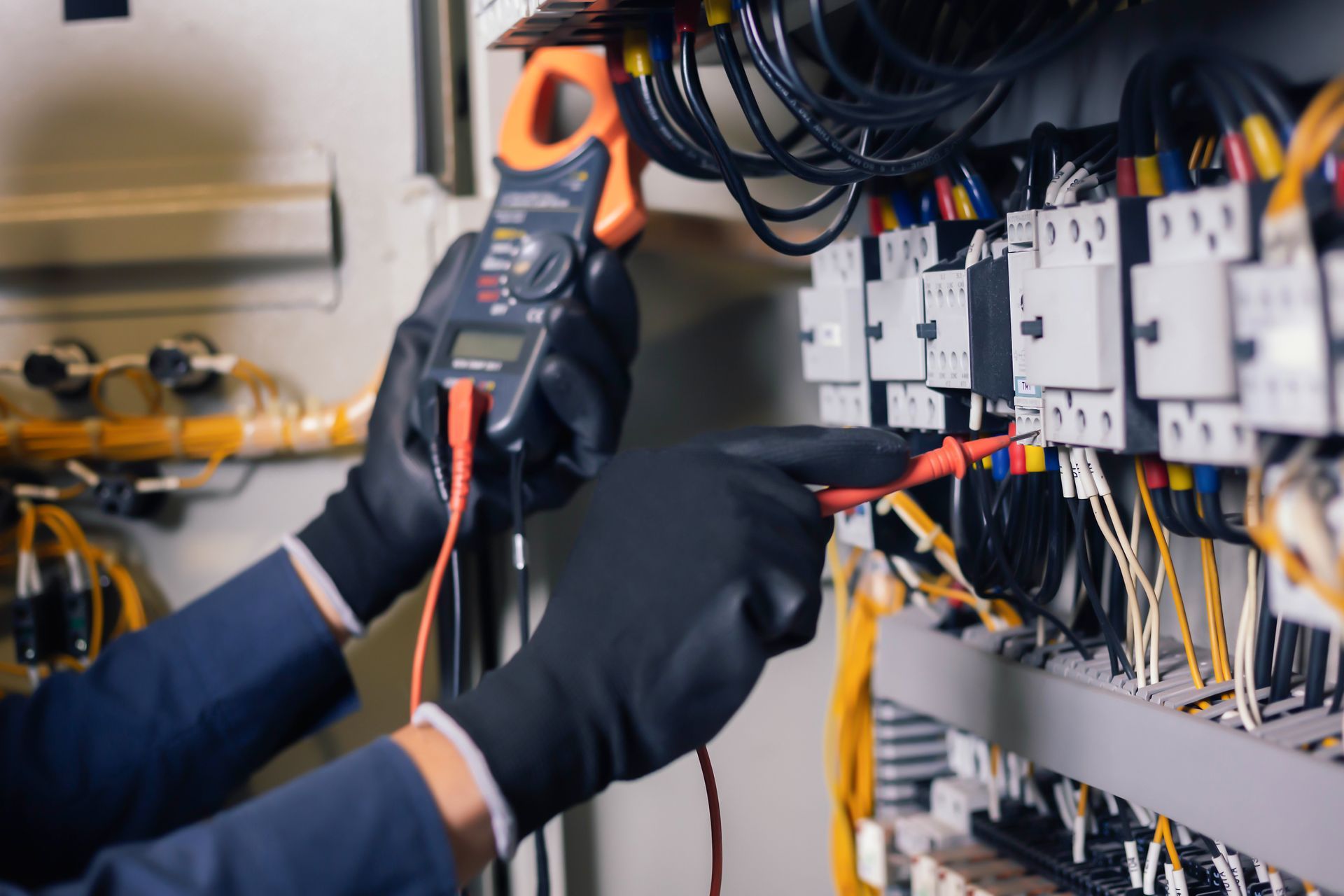Technician wearing black gloves tests wires in an electrical control panel with a multimeter.