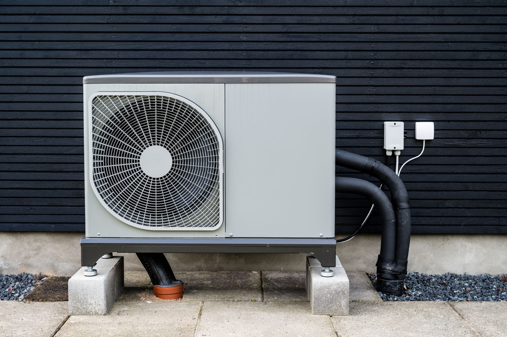 Outdoor heat pump unit against a black wall, on concrete blocks, with black pipes.