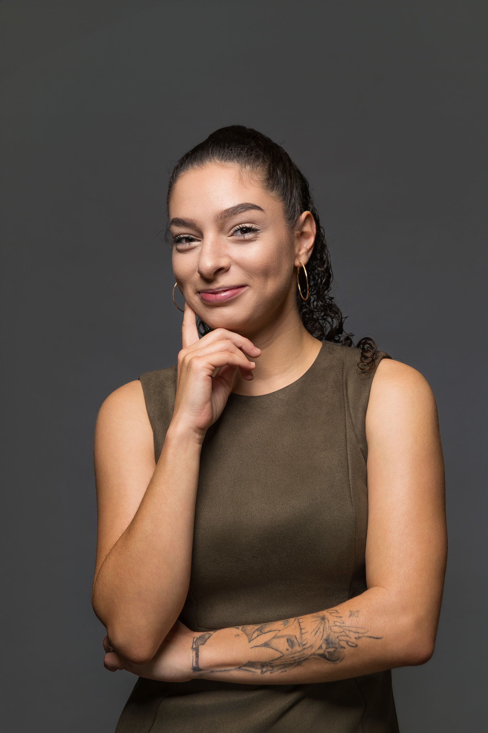 Portrait of a smiling woman in a sleeveless brown top, resting her chin on her hand against a gray background