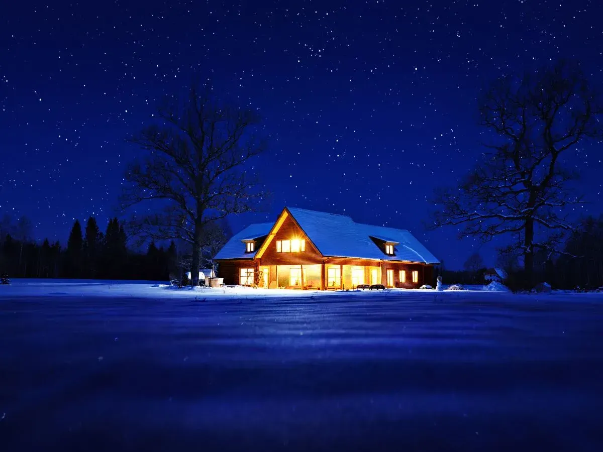 A warm, glowing house sits in a snowy landscape under a vast, star-filled night sky