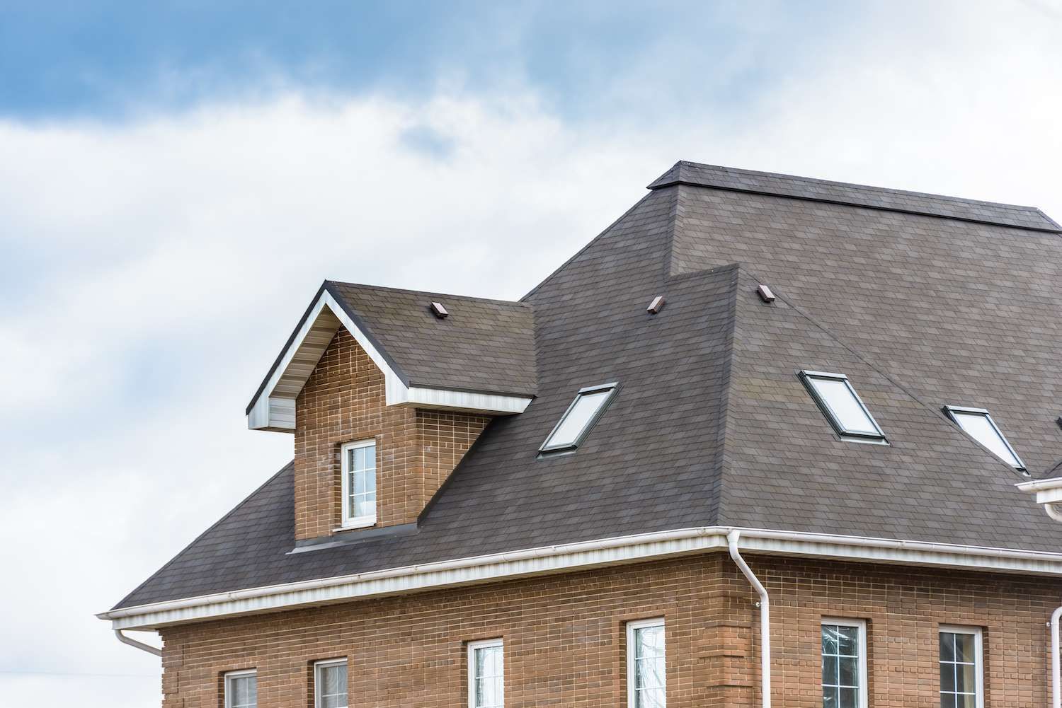 Brick building with dark shingled roof, dormer, and skylights against a cloudy sky.