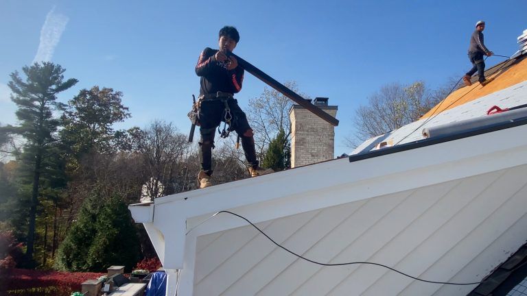 Workers on a roof with blue sky. One man carries a long, dark piece. Another man in the background.