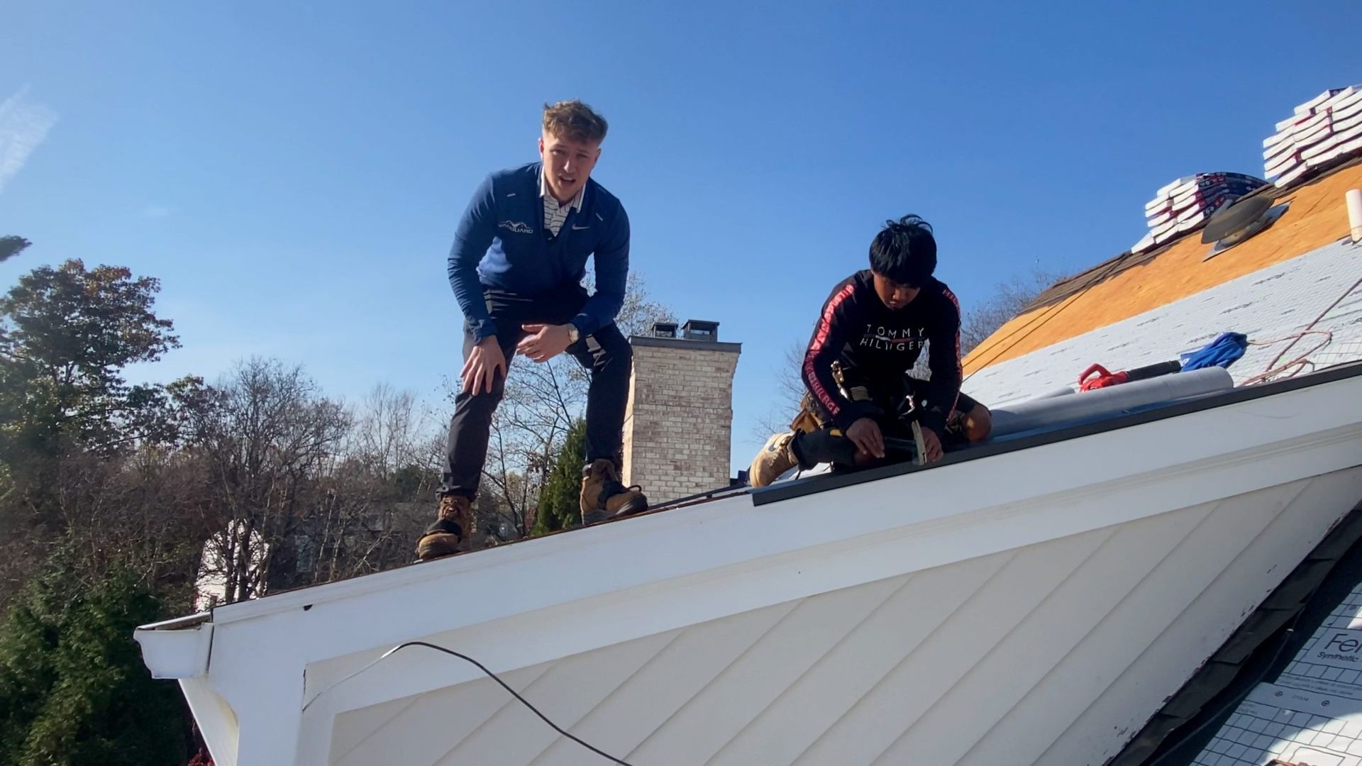 Two people working on a roof; one standing, the other crouched, under a blue sky.
