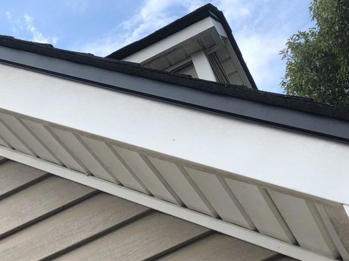 Close-up of a house roof, showing black shingles, white trim, and light brown siding against a cloudy blue sky.