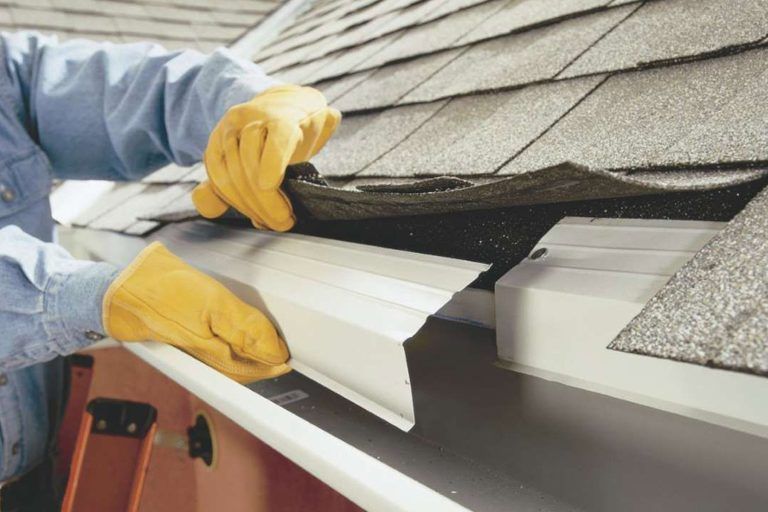 Person in yellow gloves installs a gutter guard under roof shingles.