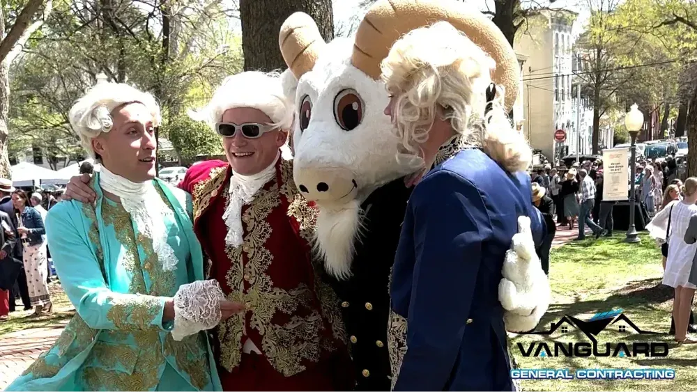 Three people in colonial costumes and a goat mascot pose for a photo outdoors.