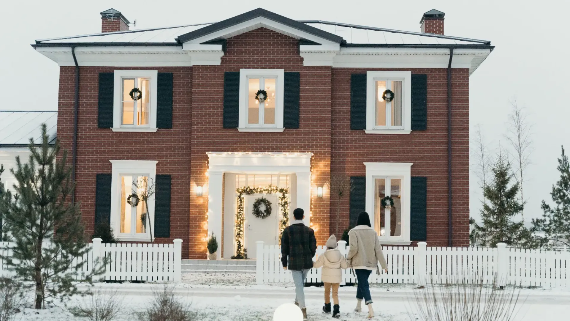 Family walking towards a festively decorated brick house with wreaths on windows and snow-covered ground.