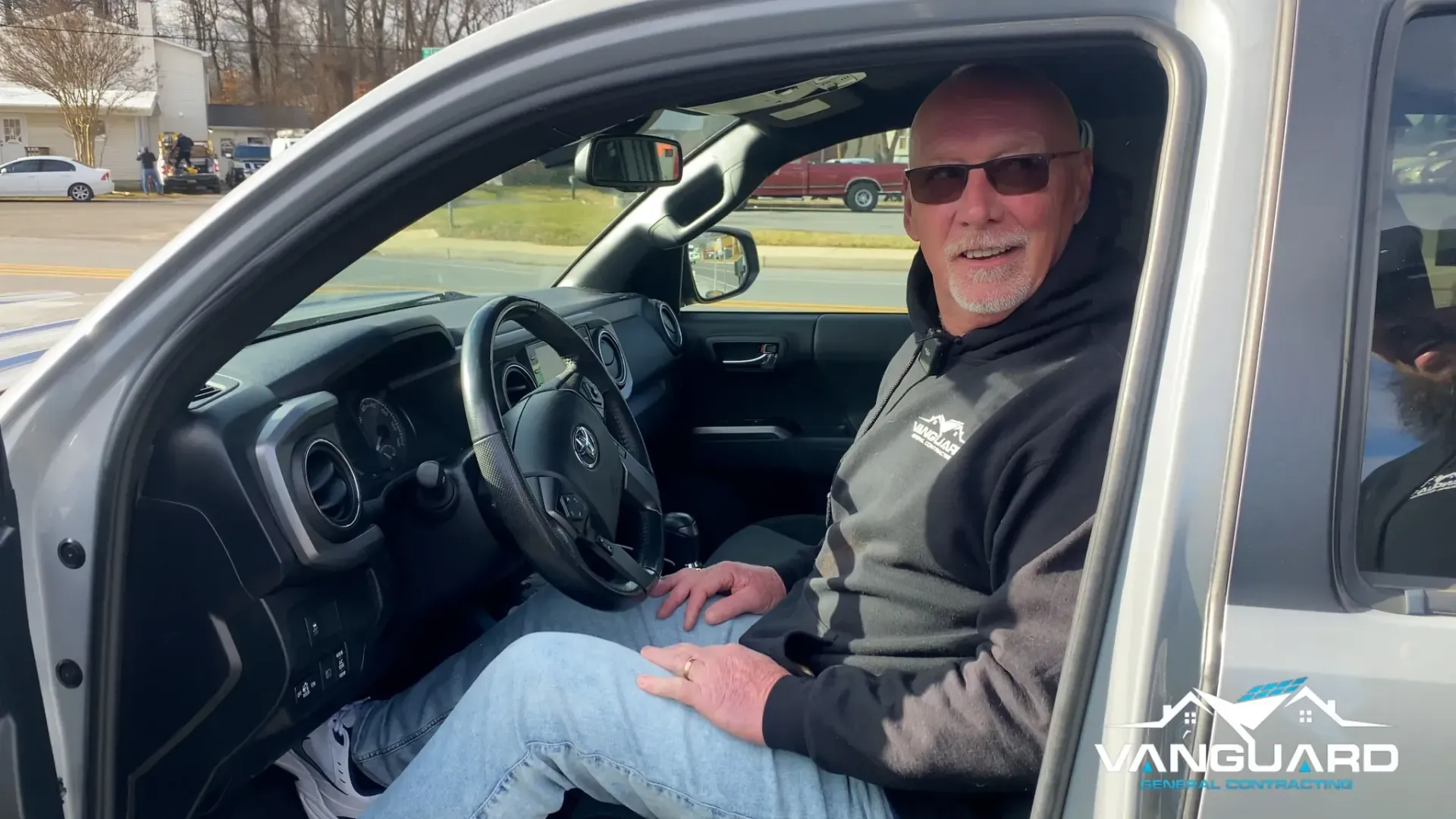 Man sitting in a truck, wearing sunglasses and a hoodie, smiling.