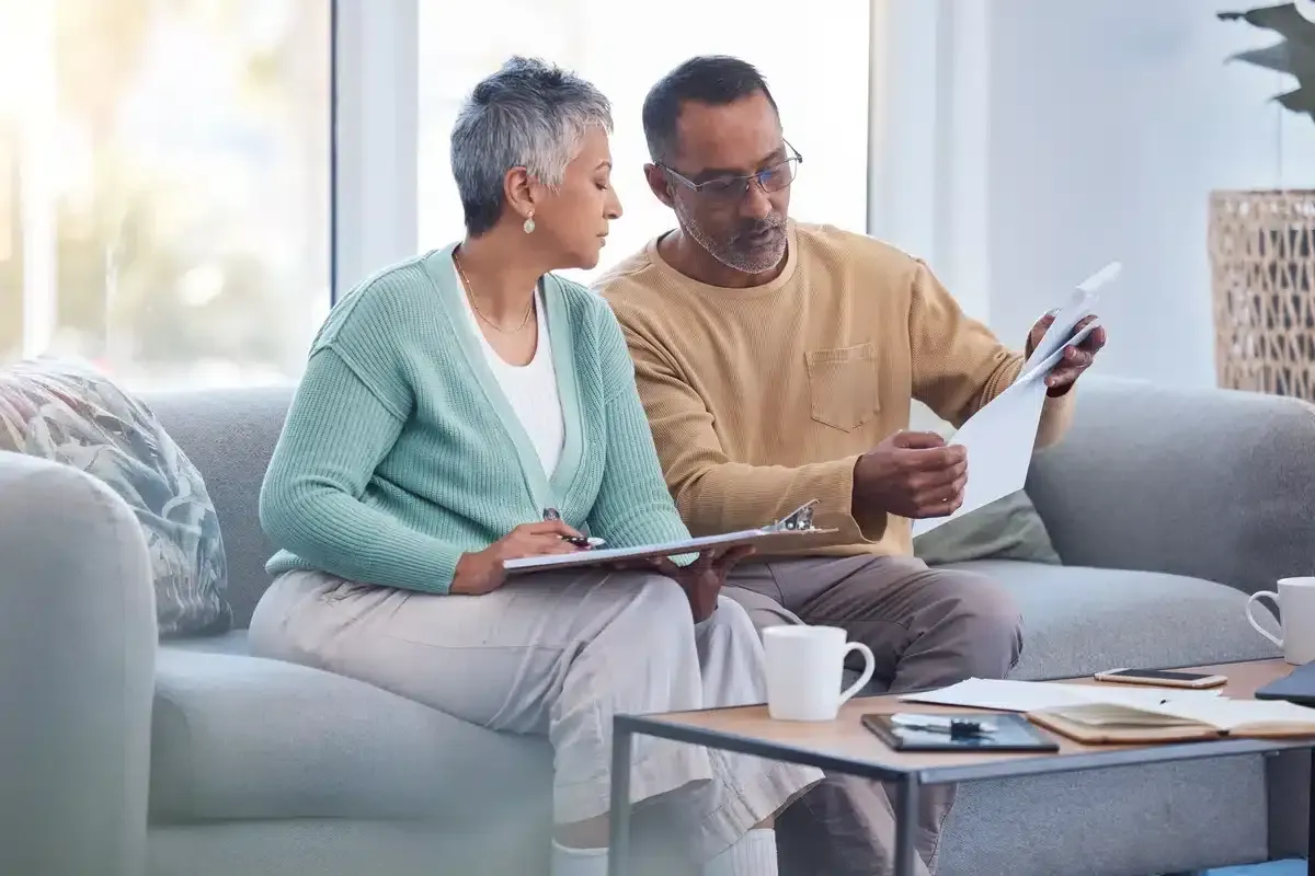 Couple reviewing documents, seated on a sofa indoors. One holds a clipboard, the other papers; both look focused.