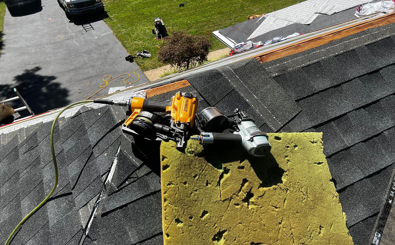 Rooftop with roofing tools: nail guns on a yellow insulation pad, near installed shingles.