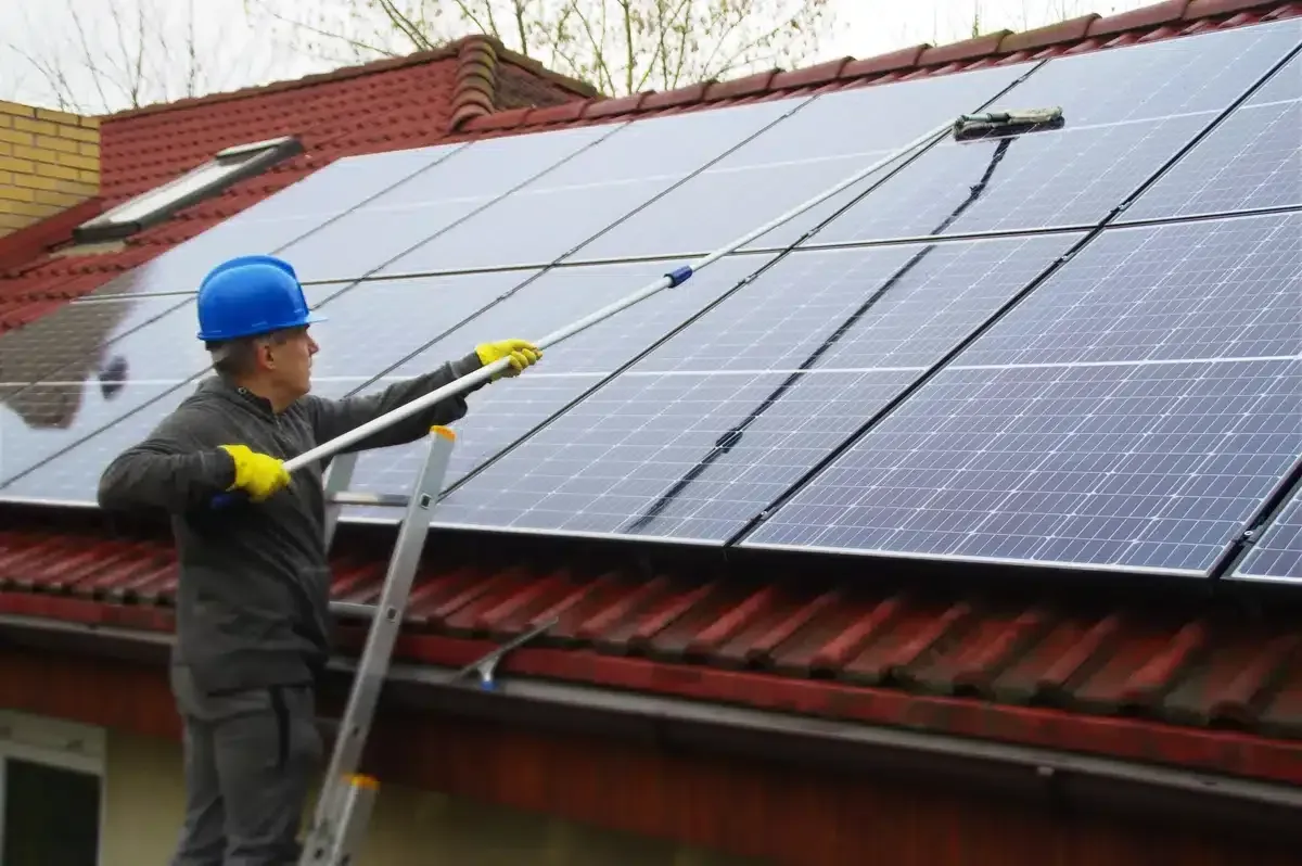 Man in blue helmet on ladder cleaning solar panels with long-handled brush on rooftop.