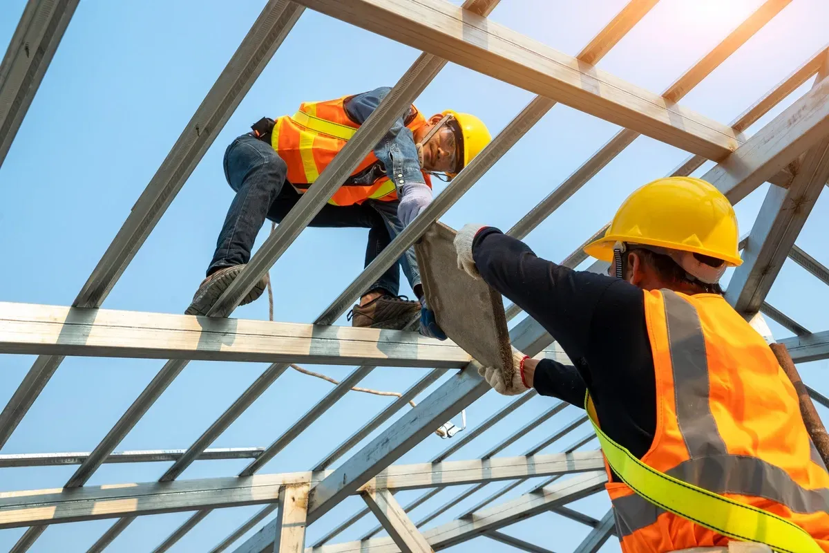 Two construction workers on a roof, one holding a tile, the other working, wearing safety vests and helmets under a blue sky.