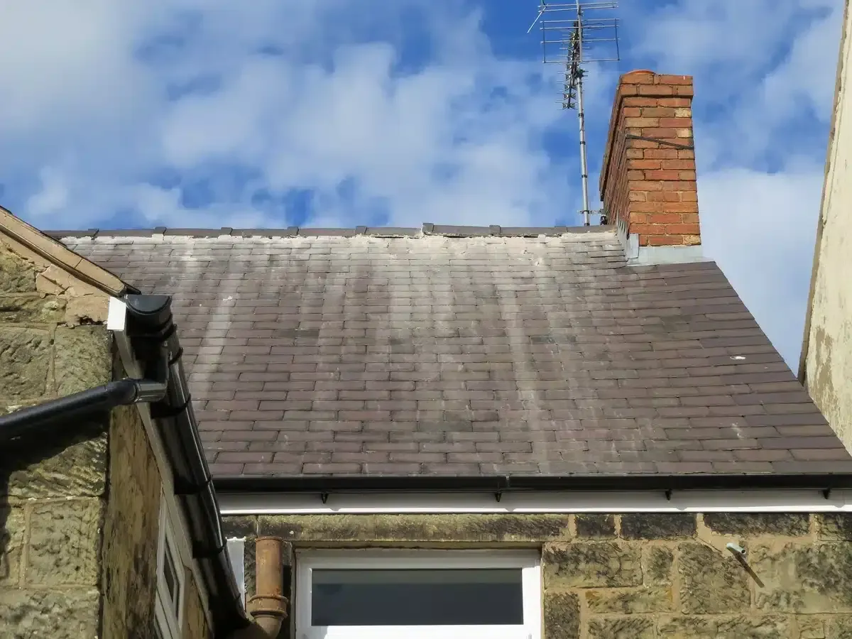 A slate roof with a brick chimney and TV antenna against a cloudy sky.