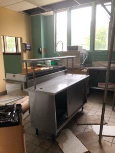 Abandoned kitchen with stainless steel counters, scattered debris, and bright windows.