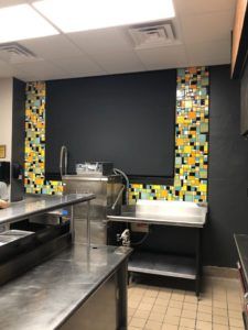Restaurant prep area with stainless steel counters, sink, and a black wall bordered by colorful mosaic tiles.