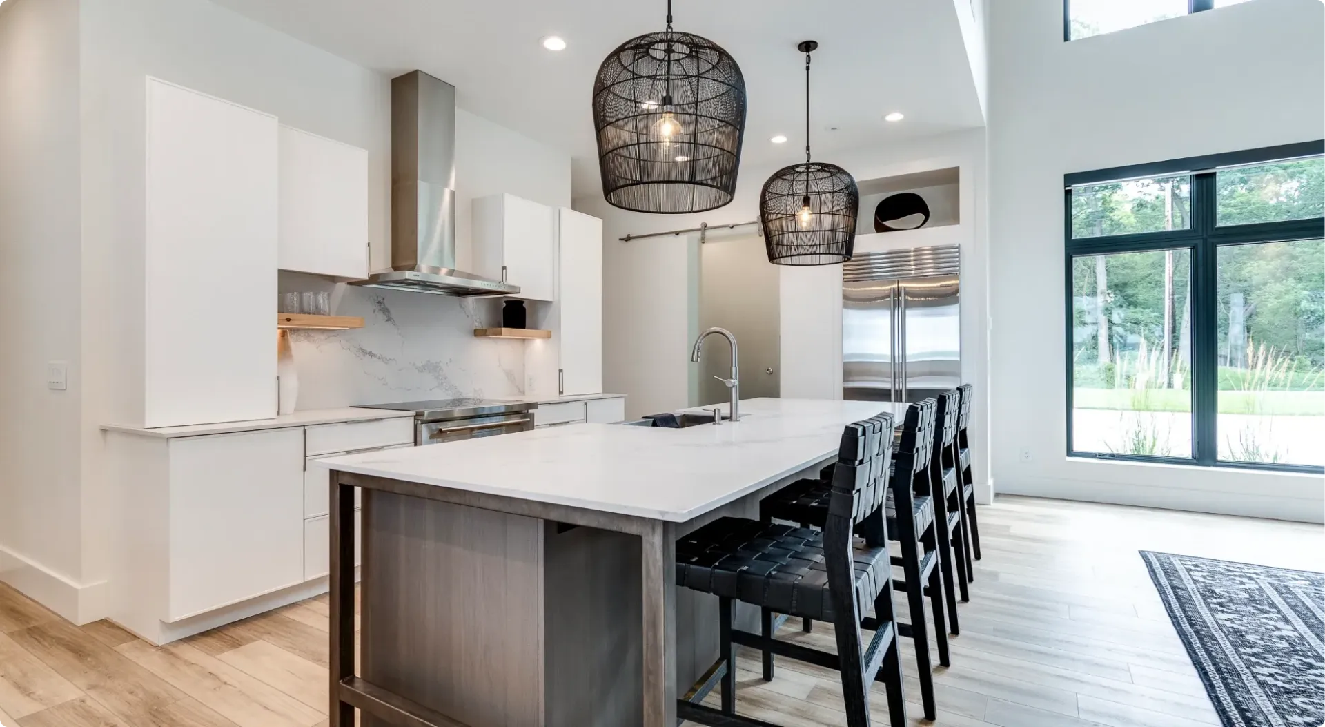 Bright modern kitchen with white cabinets, marble island, black bar stools, and large windows